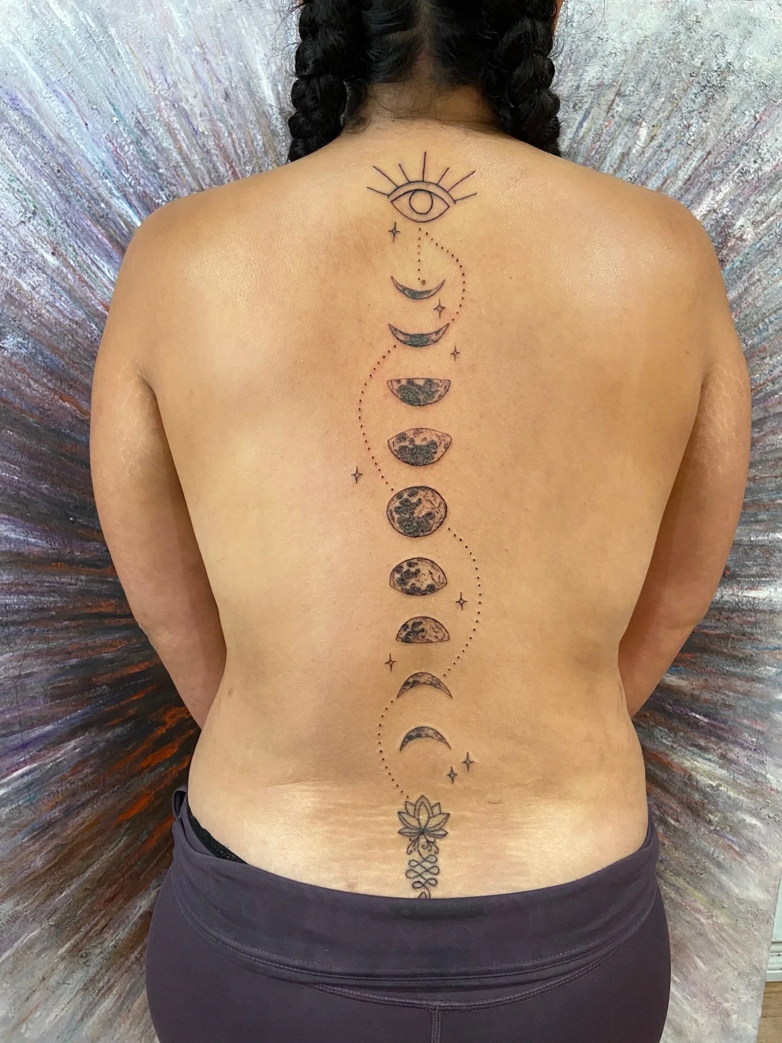 Tattoo of the moon phases, stars, an eye with rays, and a lotus flower on a person's back.