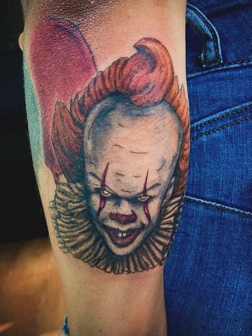 Tattoo of Pennywise the clown from It with red hair, yellow eyes, red scars, and a ruffled collar.