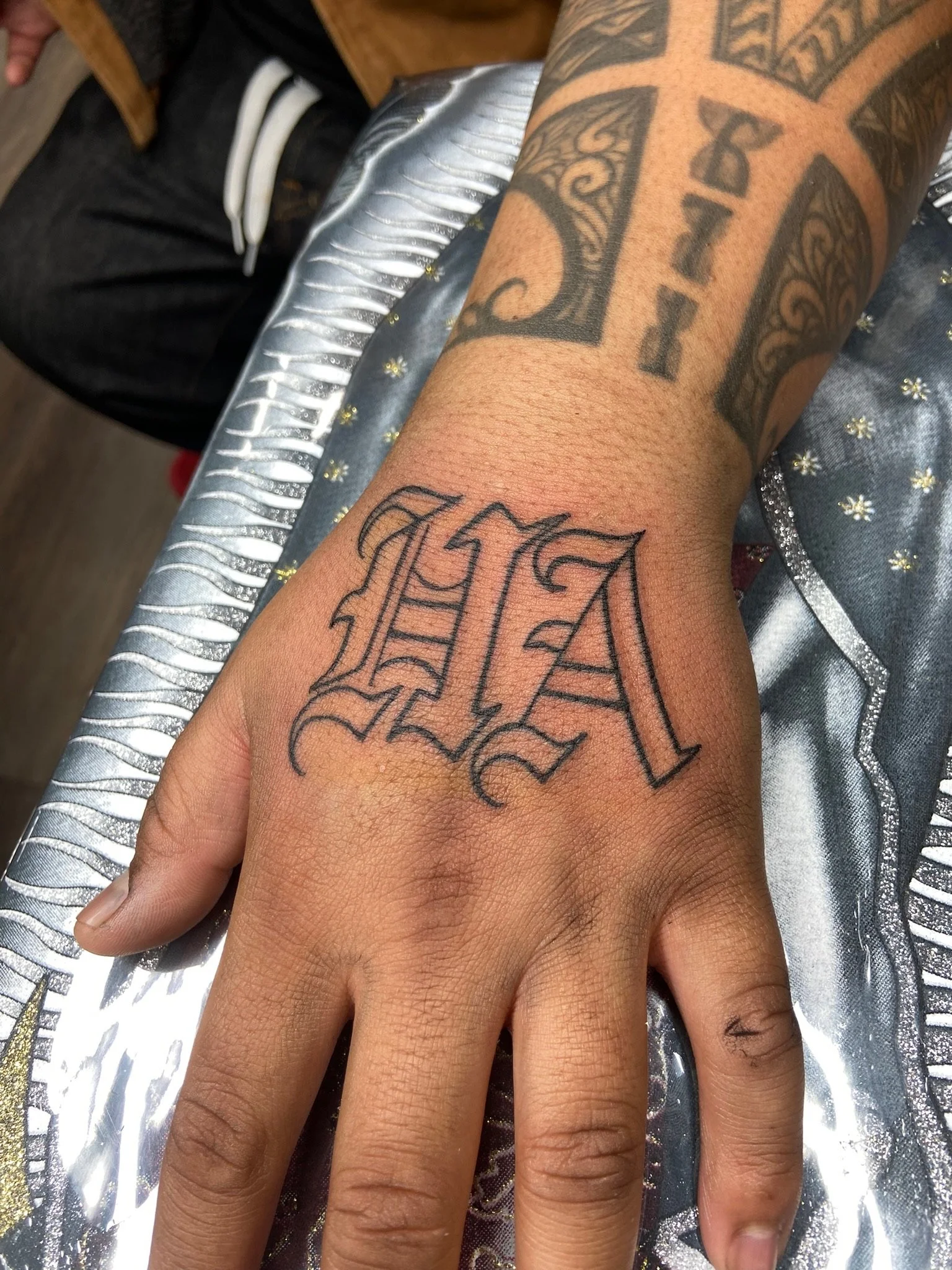 A person's hand with tattoos resting on a shiny, metallic surface with decorative accents. The tattoos include a large cross with intricate details and the word 'REAL' in stylized Gothic font.