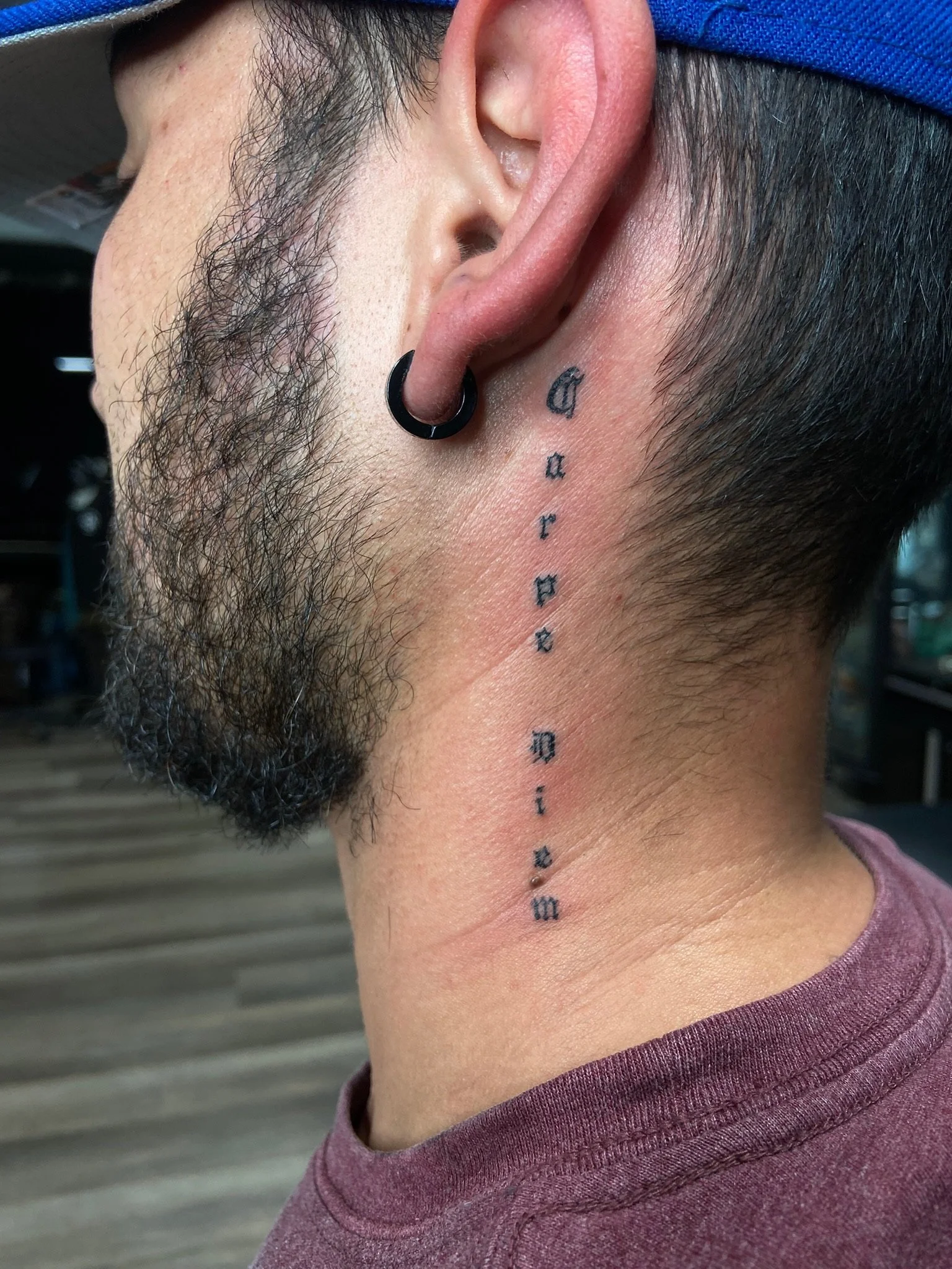 Close-up of a person's left side head showing facial hair, a newly pierced ear with a black hoop earring, and a tattoo of small, black letters or symbols running down the neck.