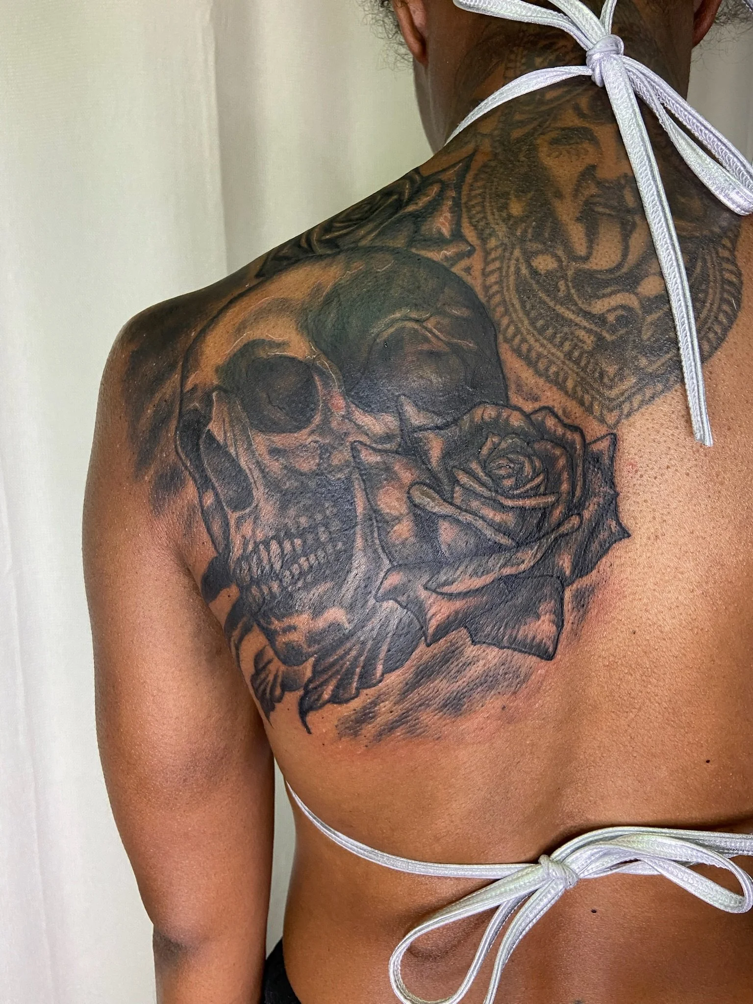 A person with a large tattoo of skulls, roses, and decorative designs on their upper arm and shoulder, wearing a white ribbon tied around their neck.