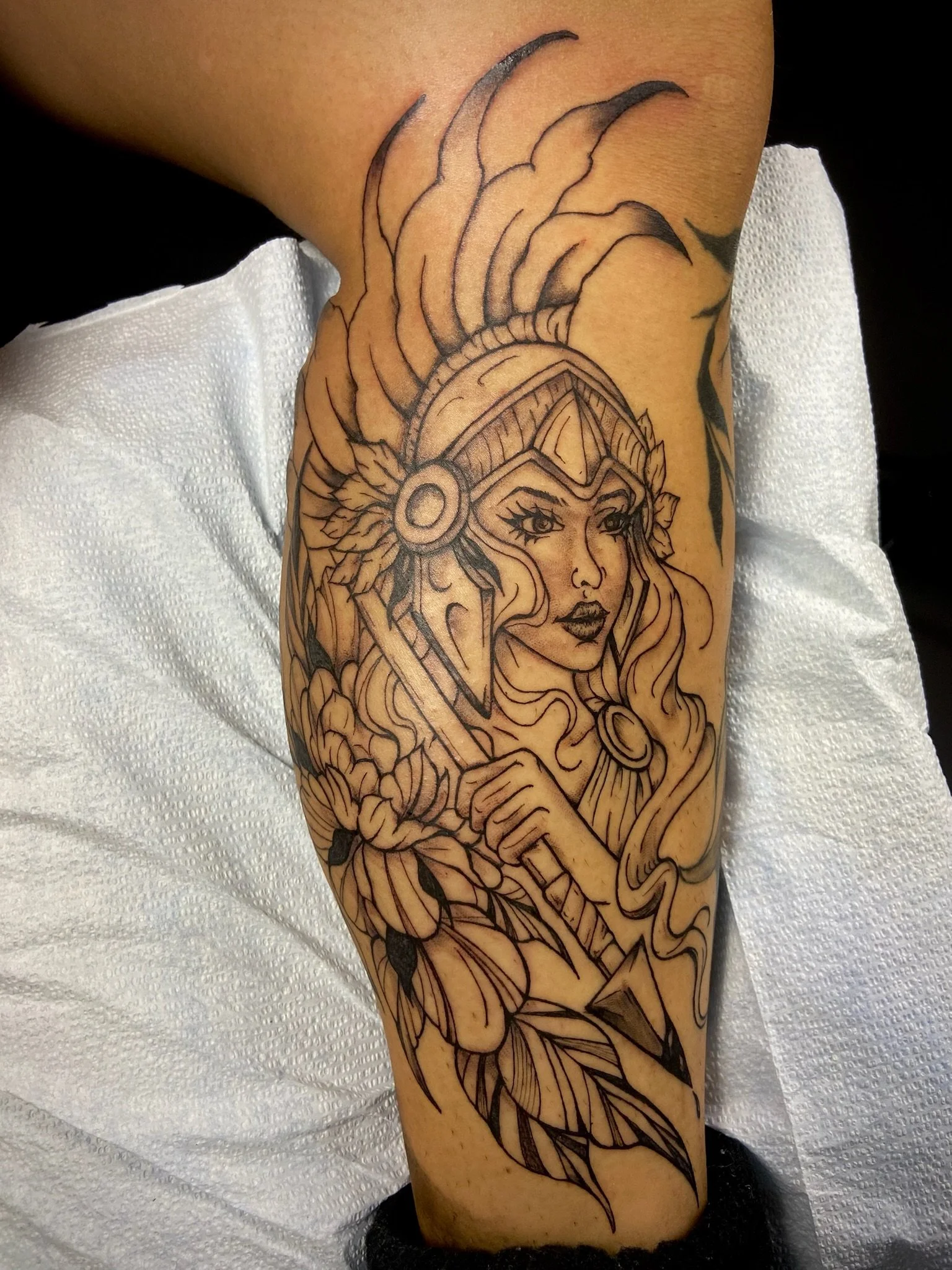 Black outline tattoo of a woman wearing a helmet with feathers, holding a spear, on a person's arm.