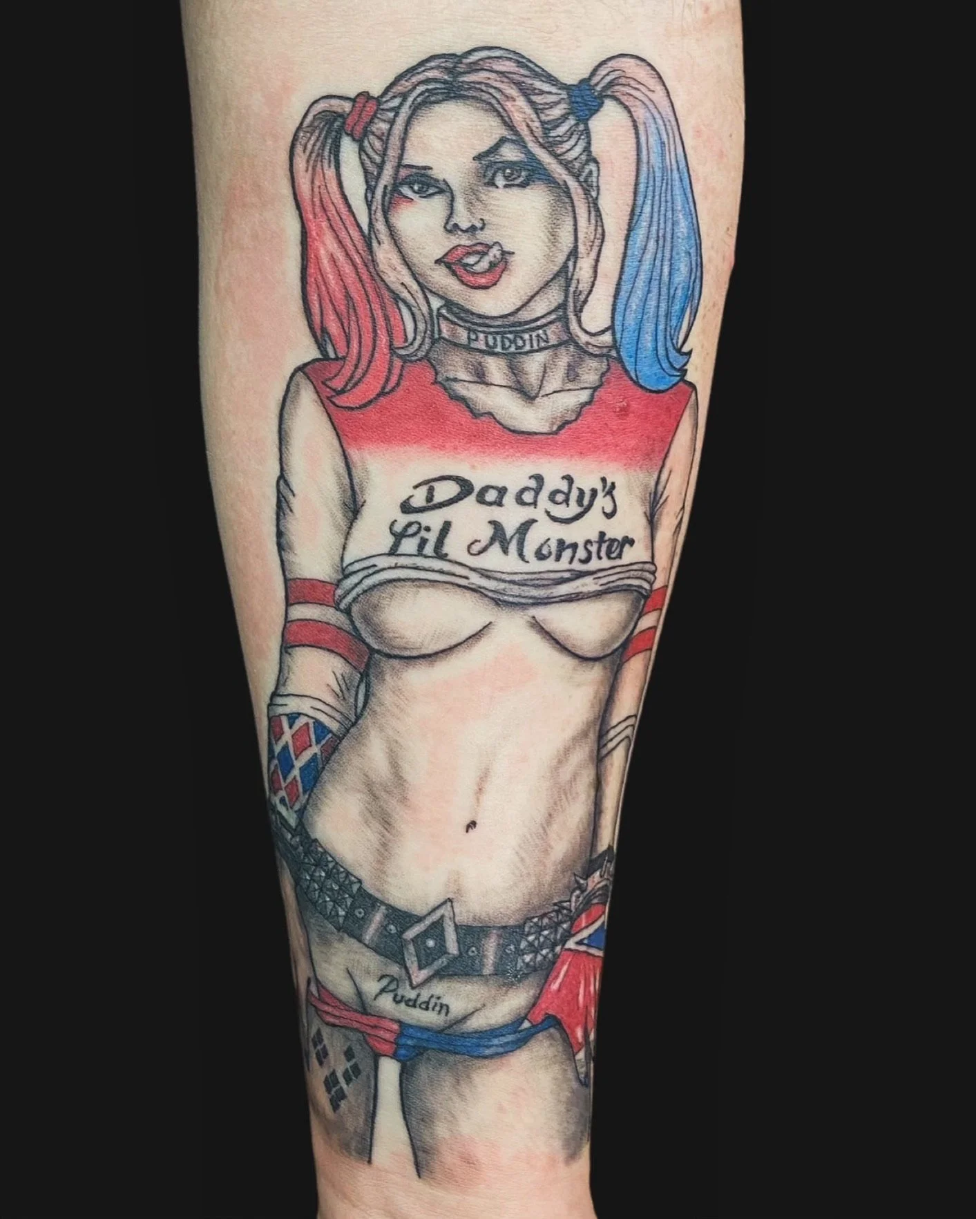 Tattoo of a woman with colorful hair, punk style clothing, and the words "Daddy's Lil Monster" on her shirt, resembling Harley Quinn from DC Comics.