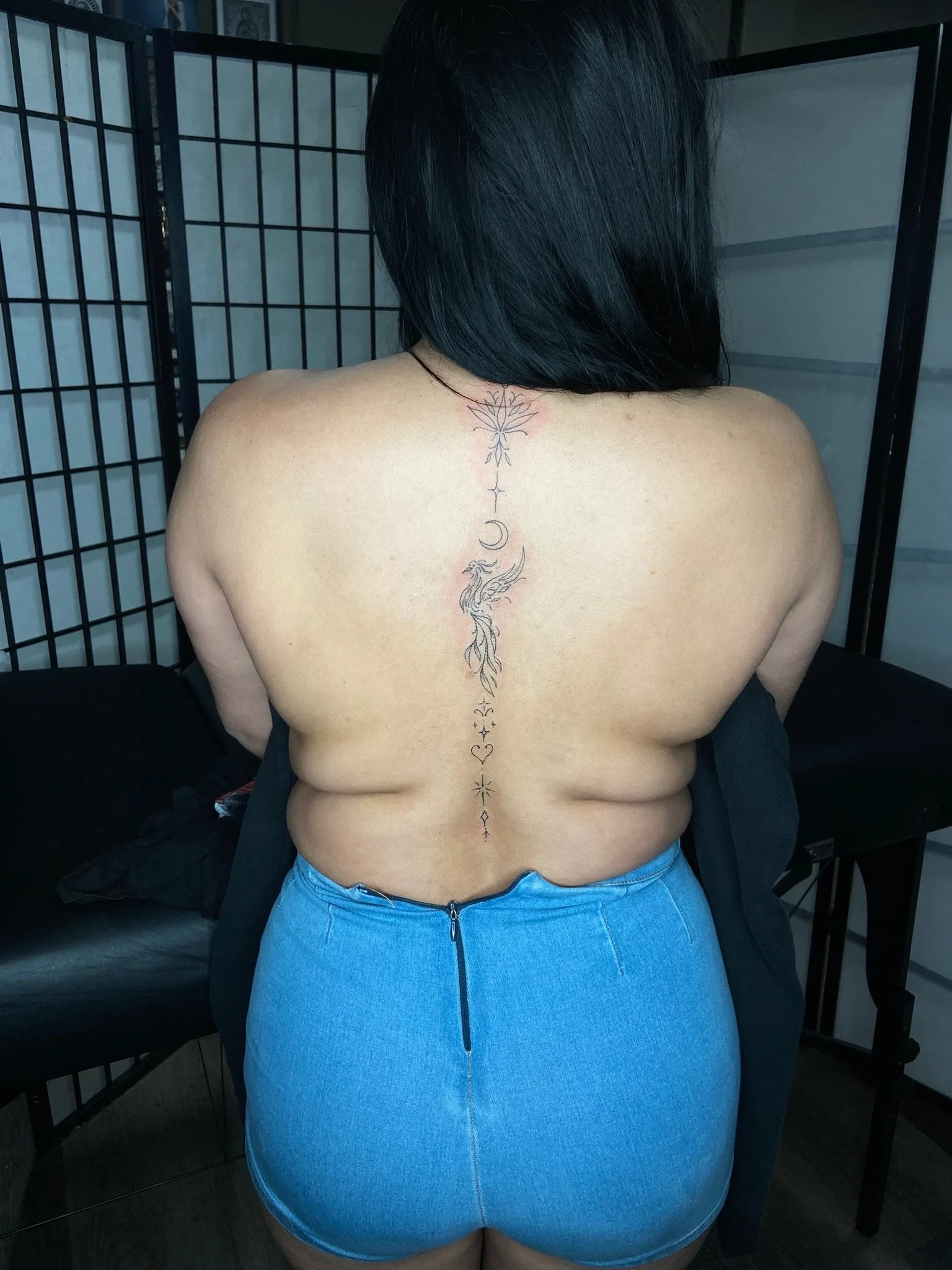 Back of a person with black hair and a tattoo of a moon, stars, and a phoenix on their upper back, wearing blue jeans.