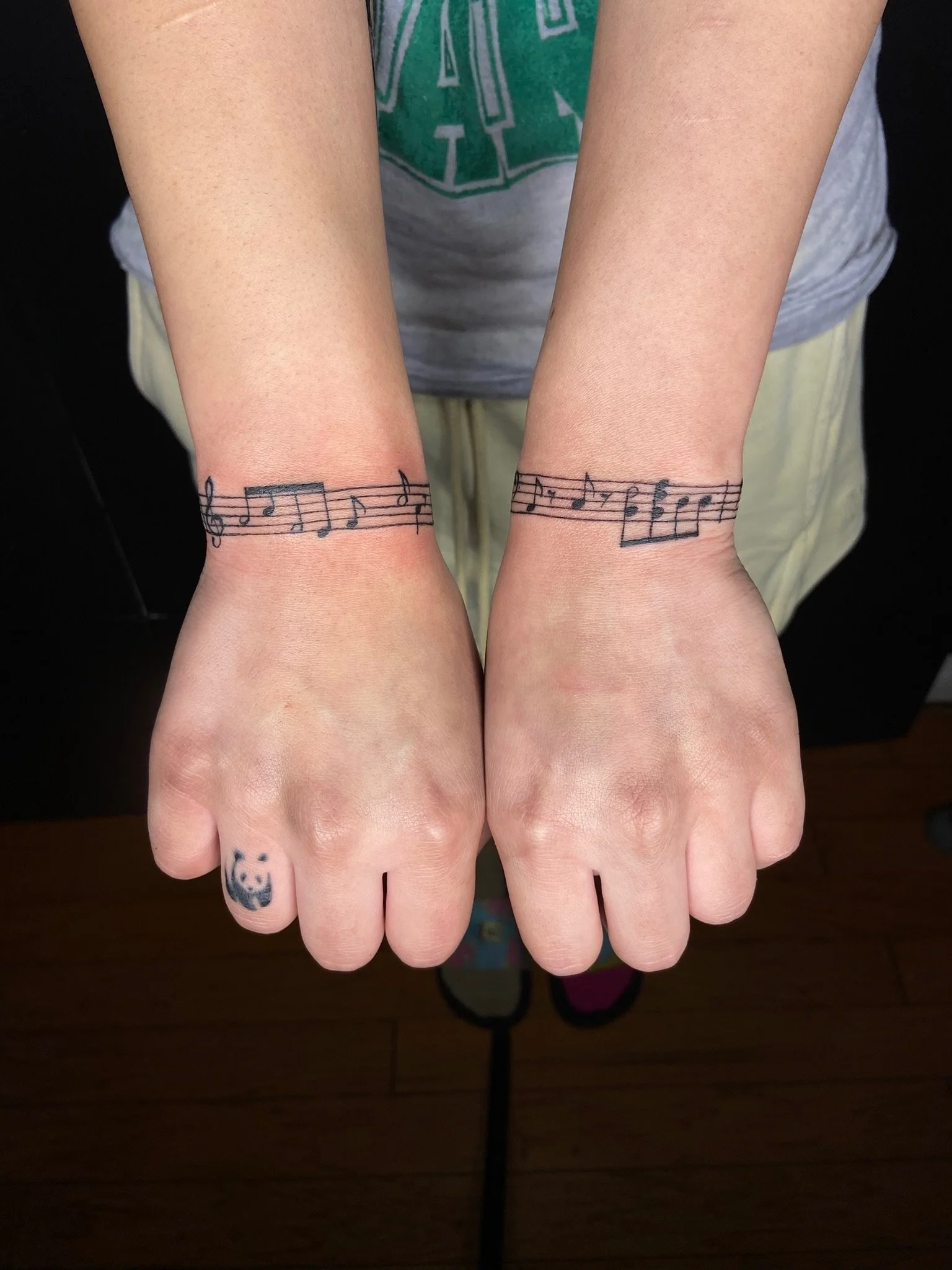 Person with small tattoos of music notes on wrists and panda face on right middle finger, wearing a gray t-shirt with green design, standing on a wooden floor.