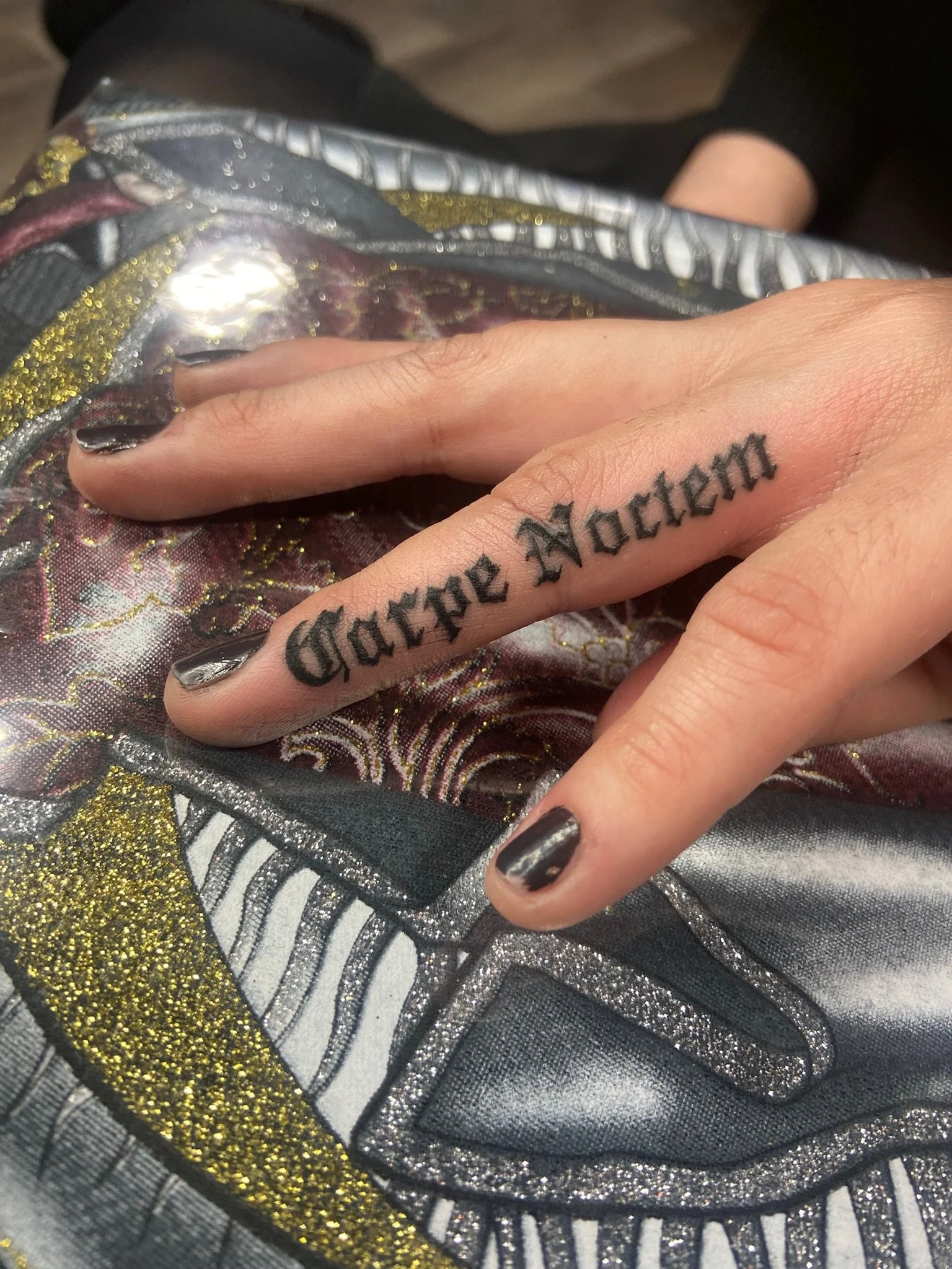 A hand with black painted nails and a tattoo that reads 'GROOVE MOTHER' resting on a fabric with gold, silver, and black intricate designs.