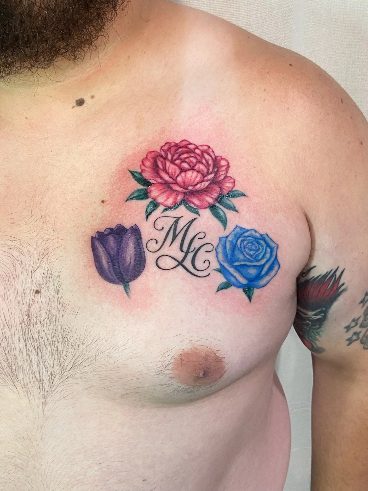 Close-up of a man's upper chest, showing tattoos including a pink-peony flower, a purple tulip, and a blue rose, with a beard visible in the top left corner.