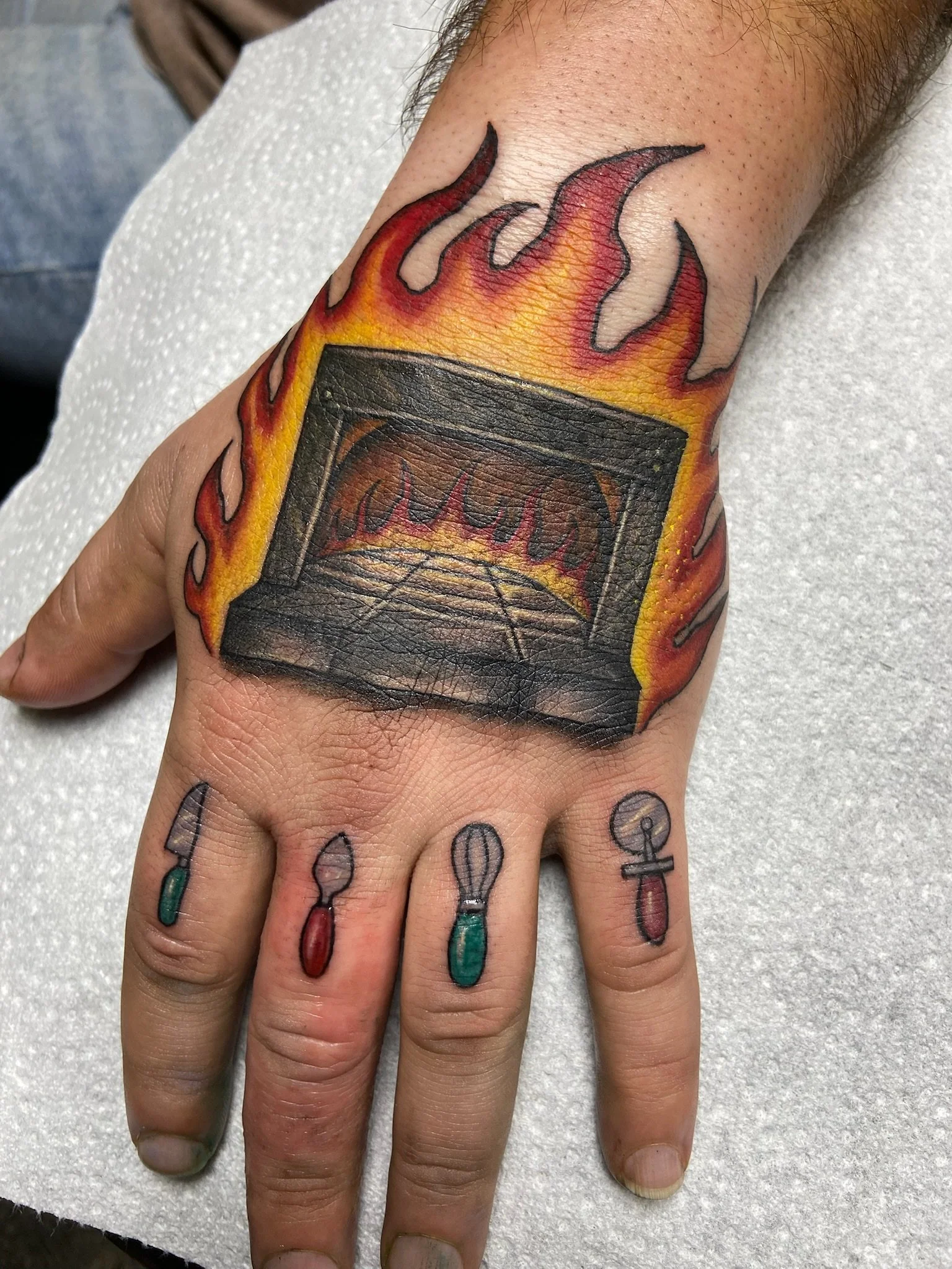A person's hand with a tattoo of a black rectangular object surrounded by flames on the top. The person has smaller tattooed items on the fingers, including colorful ink and symbols.