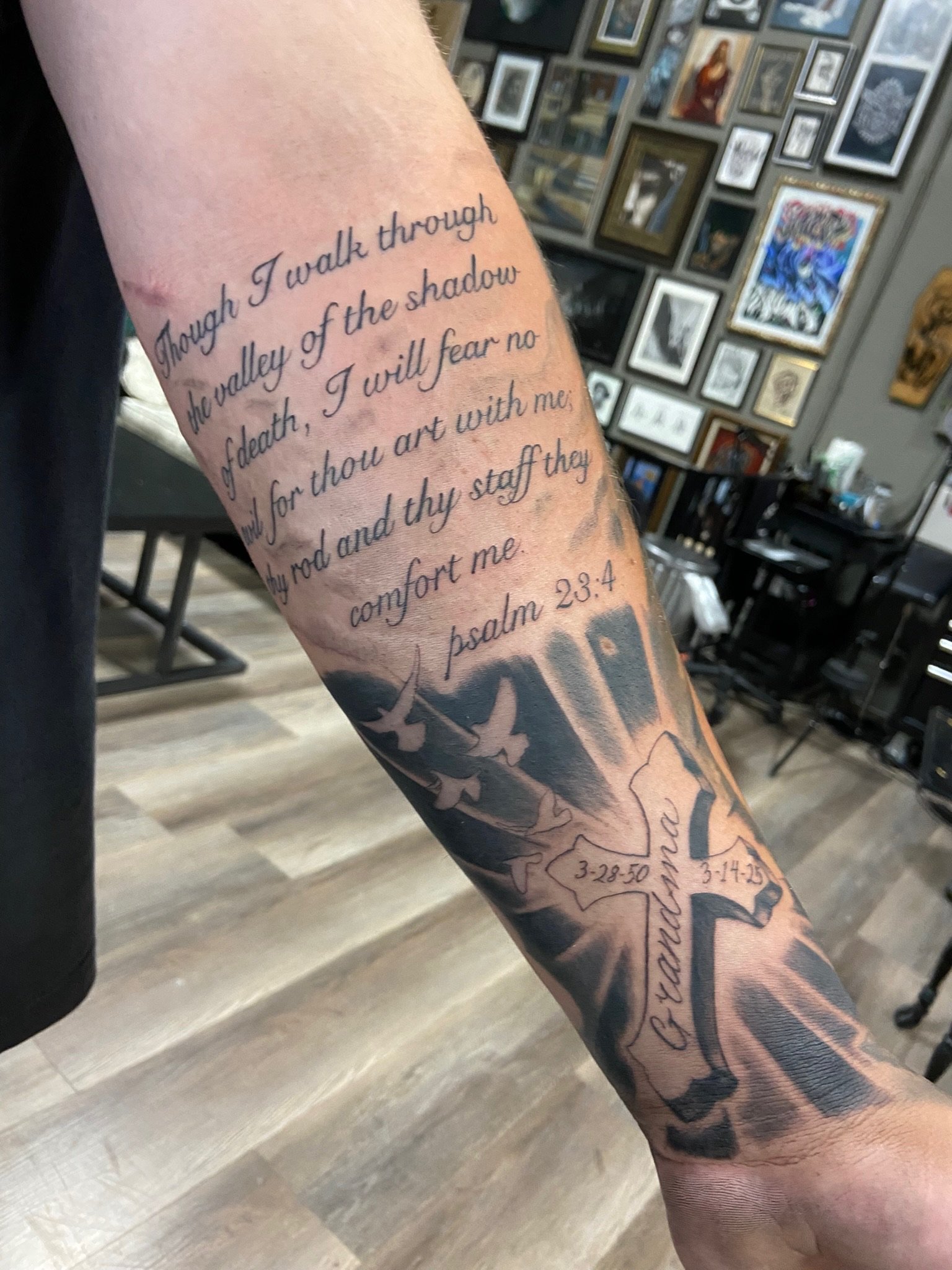 Tattoo on arm with a scripture from Psalm 23:4, surrounded by tattooed mountains and dates, inside a room with framed artwork on the wall.