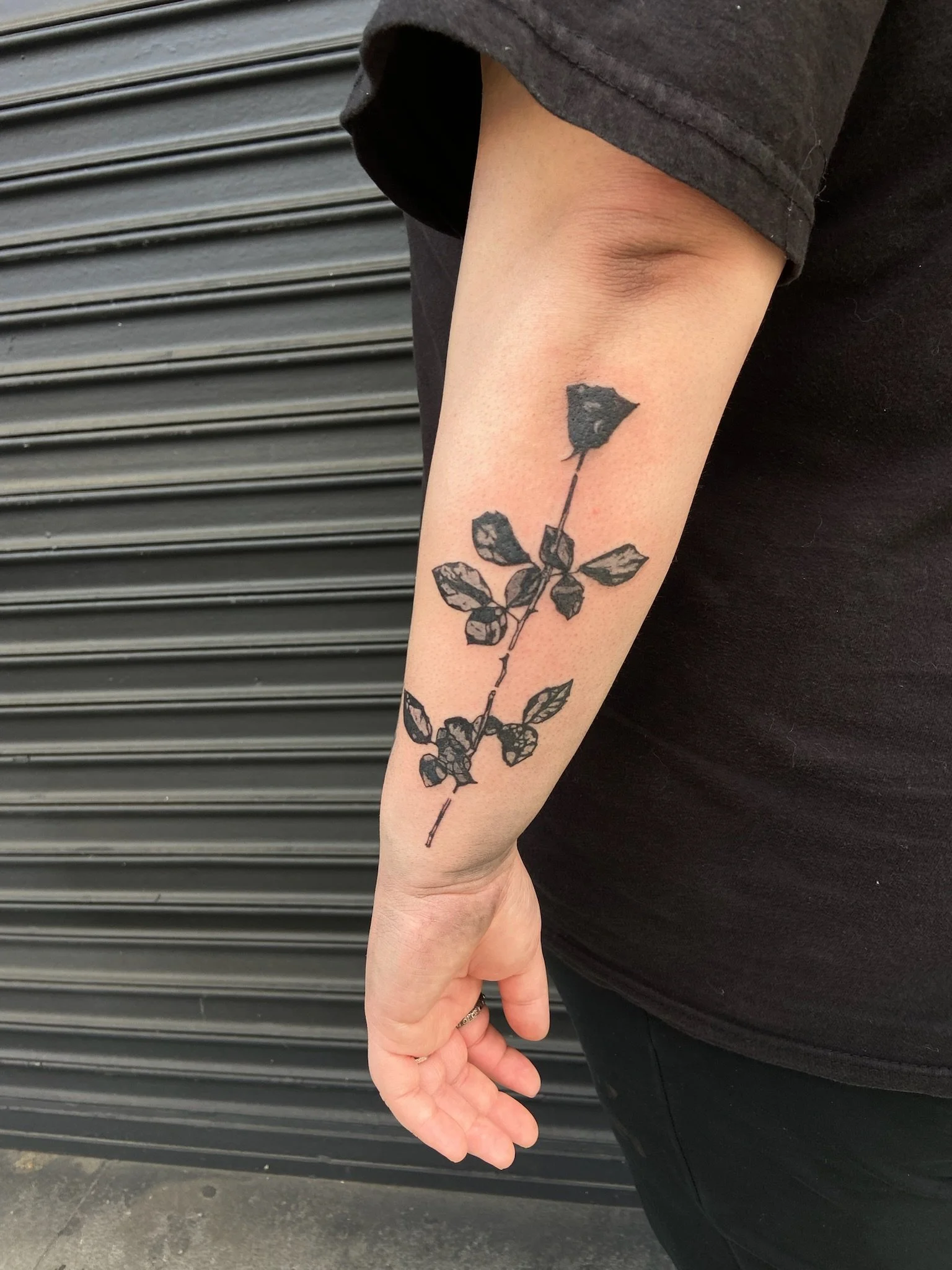 A person's forearm with a black ink tattoo of a rose with leaves. The person is wearing a dark shirt and black pants, standing in front of a metal roll-up door.