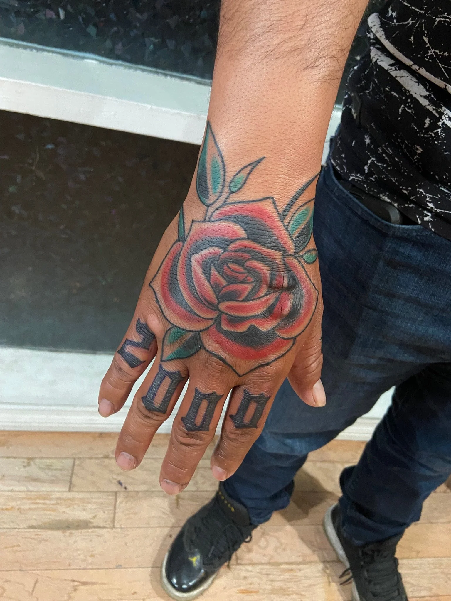 A person's hand with a colorful rose tattoo on the wrist and fingers, wearing black sneakers with yellow accents, and blue jeans, standing on a wooden floor.