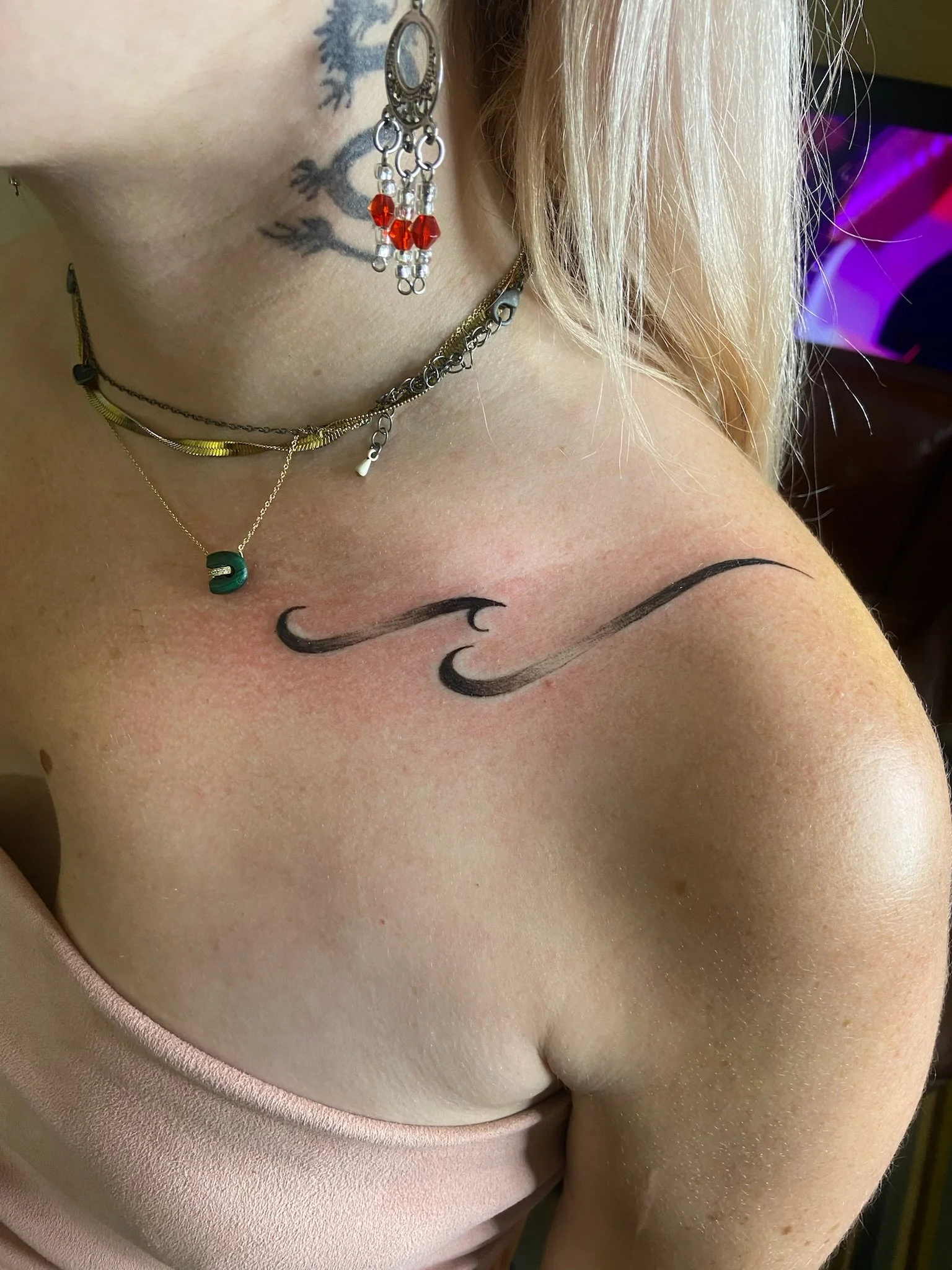 Close-up of a woman's shoulder showing jewelry necklaces, earrings, tattoos of two black curved lines, and part of a pink top with a beige background.