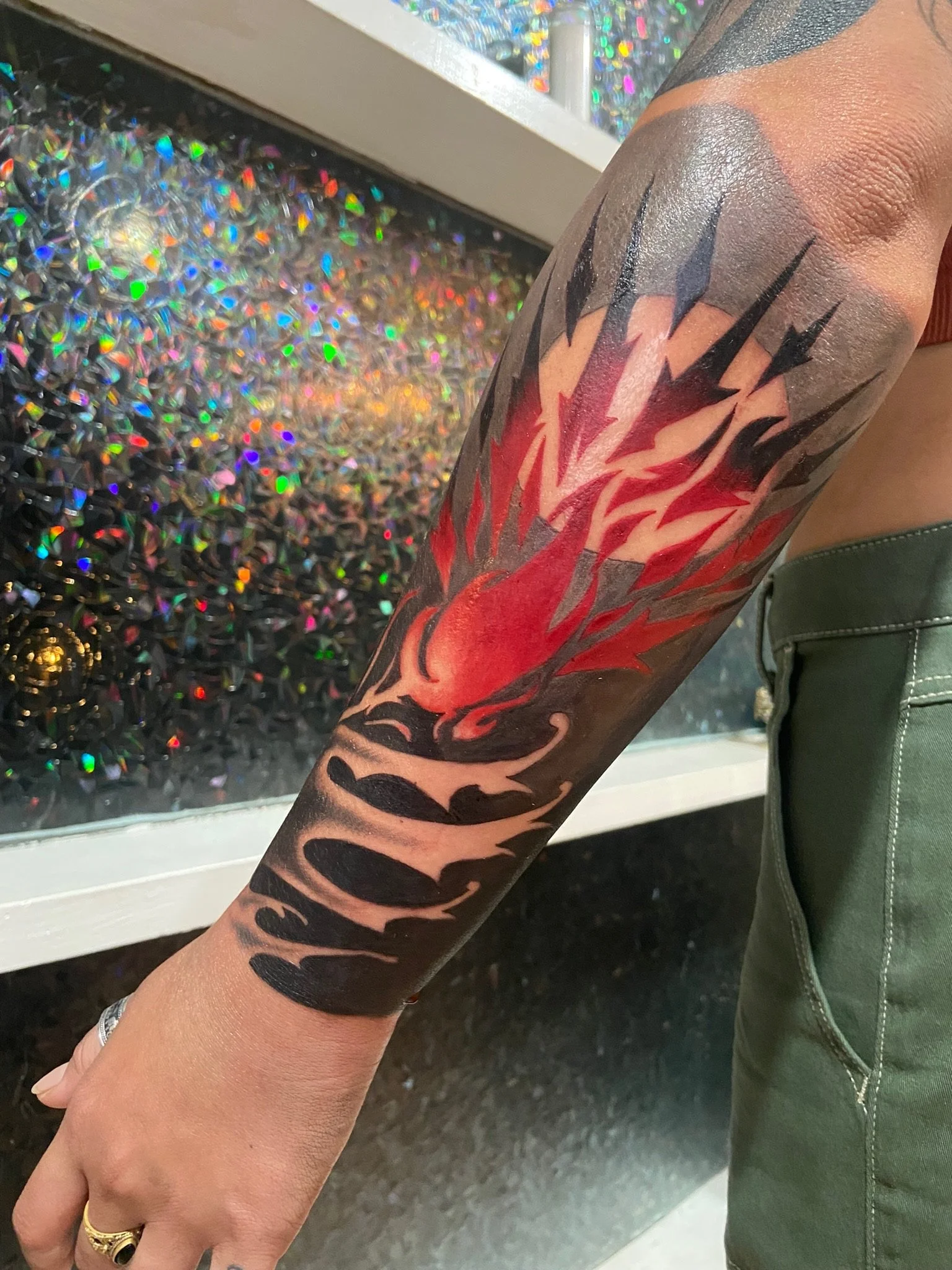 Close-up of a person’s arm with a colorful tattoo of red and black flames, showing intricate details and shading, with a background of holographic, iridescent decoration.