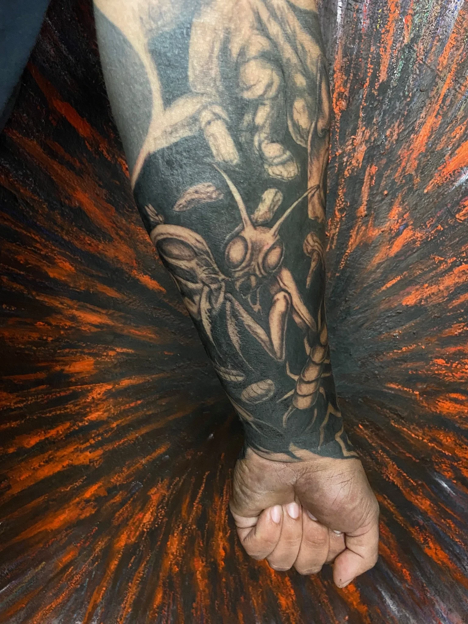 A person's hand holding a tattooed arm, with a colorful, painted background resembling a swirling, fiery pattern.