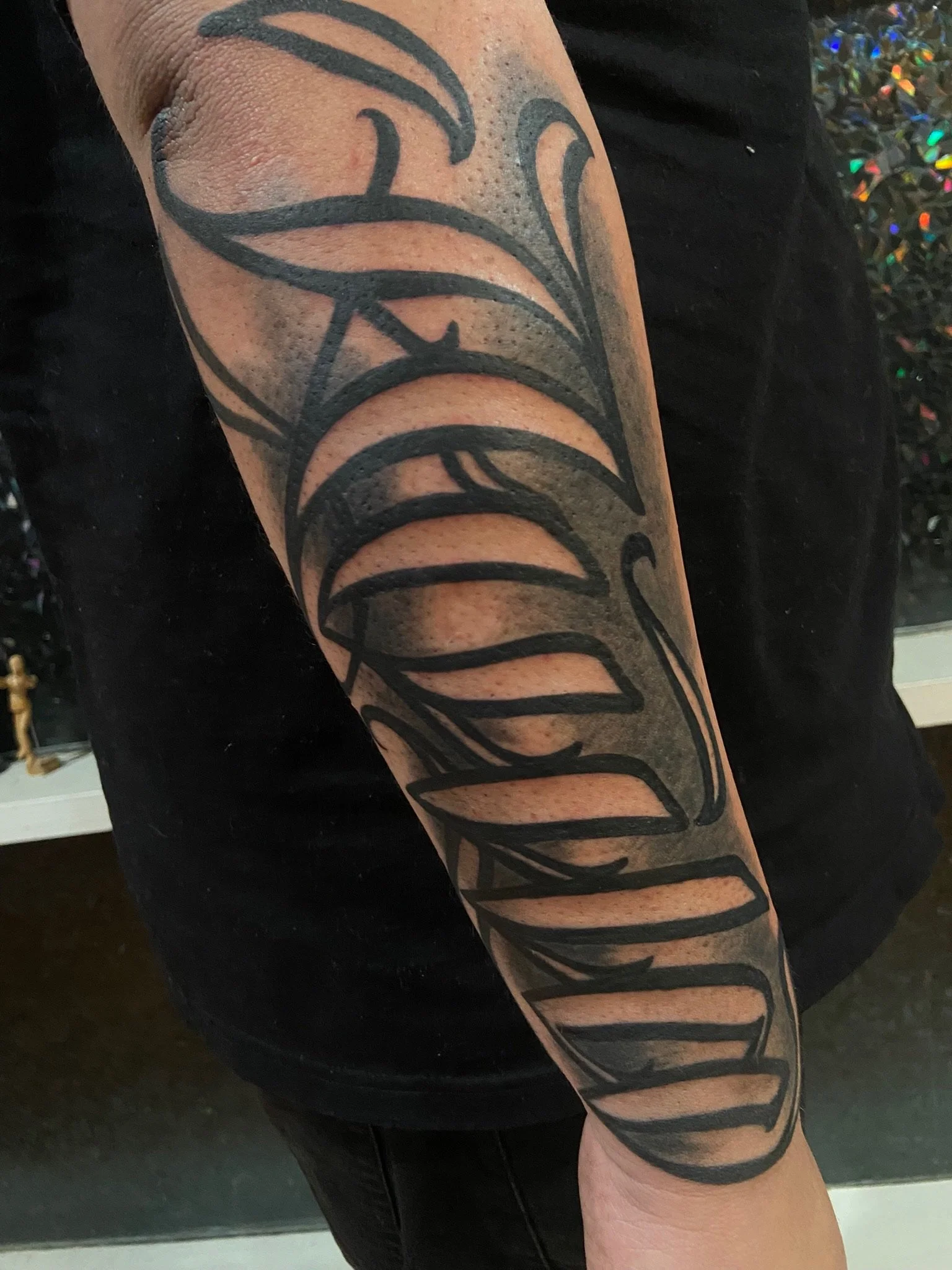 A person's arm with a large black tattoo featuring bold, flowing script and abstract lines.