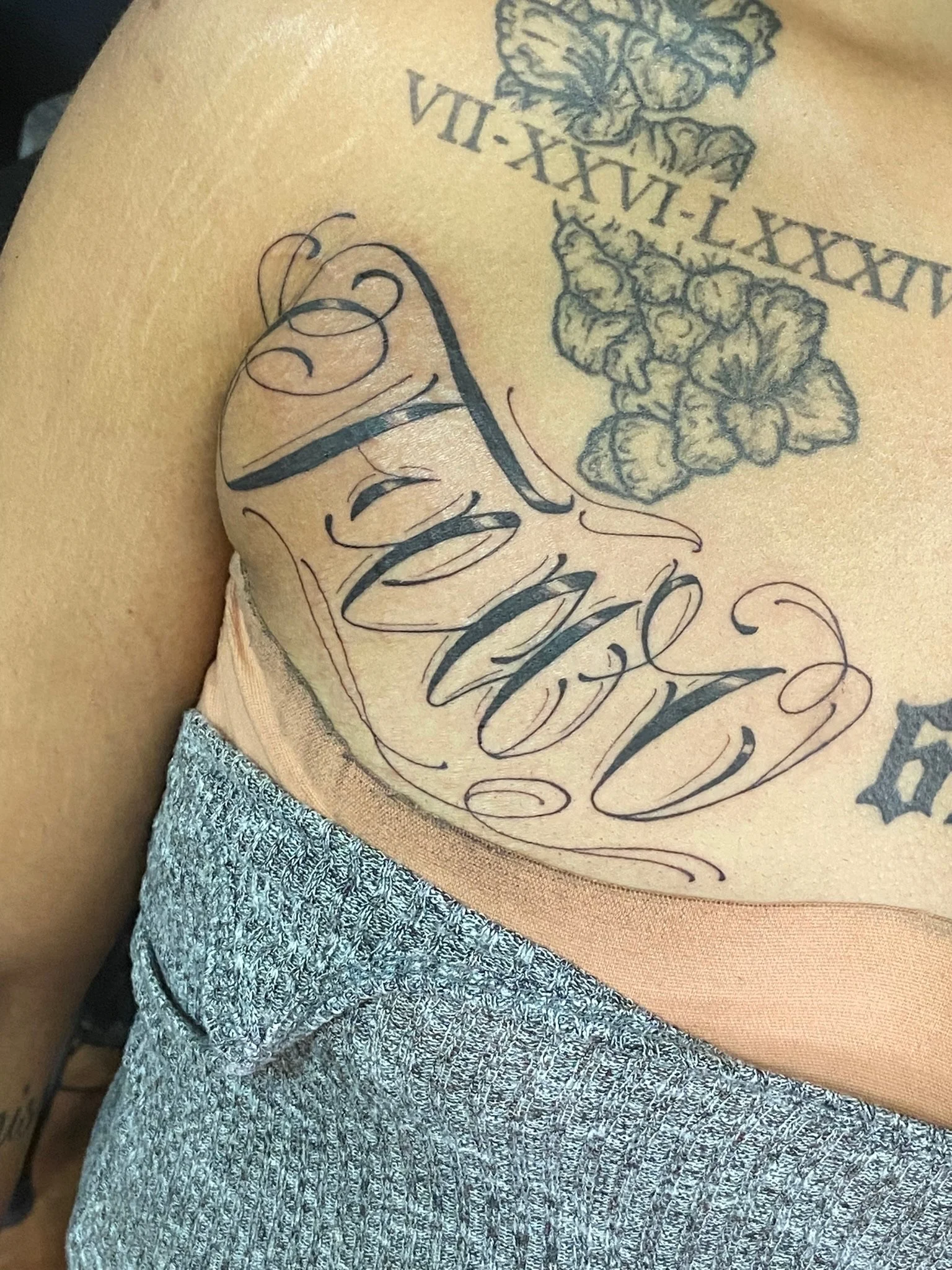 Close-up of a person's upper chest and shoulder area with multiple tattoos, including a floral design, a phrase in cursive, and a crown.