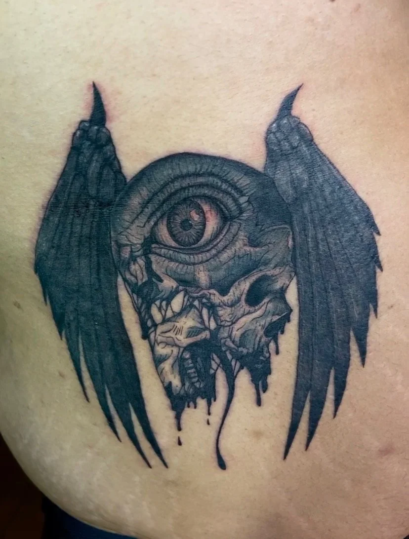 Tattoo of a creature with a large single eye, sharp teeth, horns, and black wings, with dripping details.