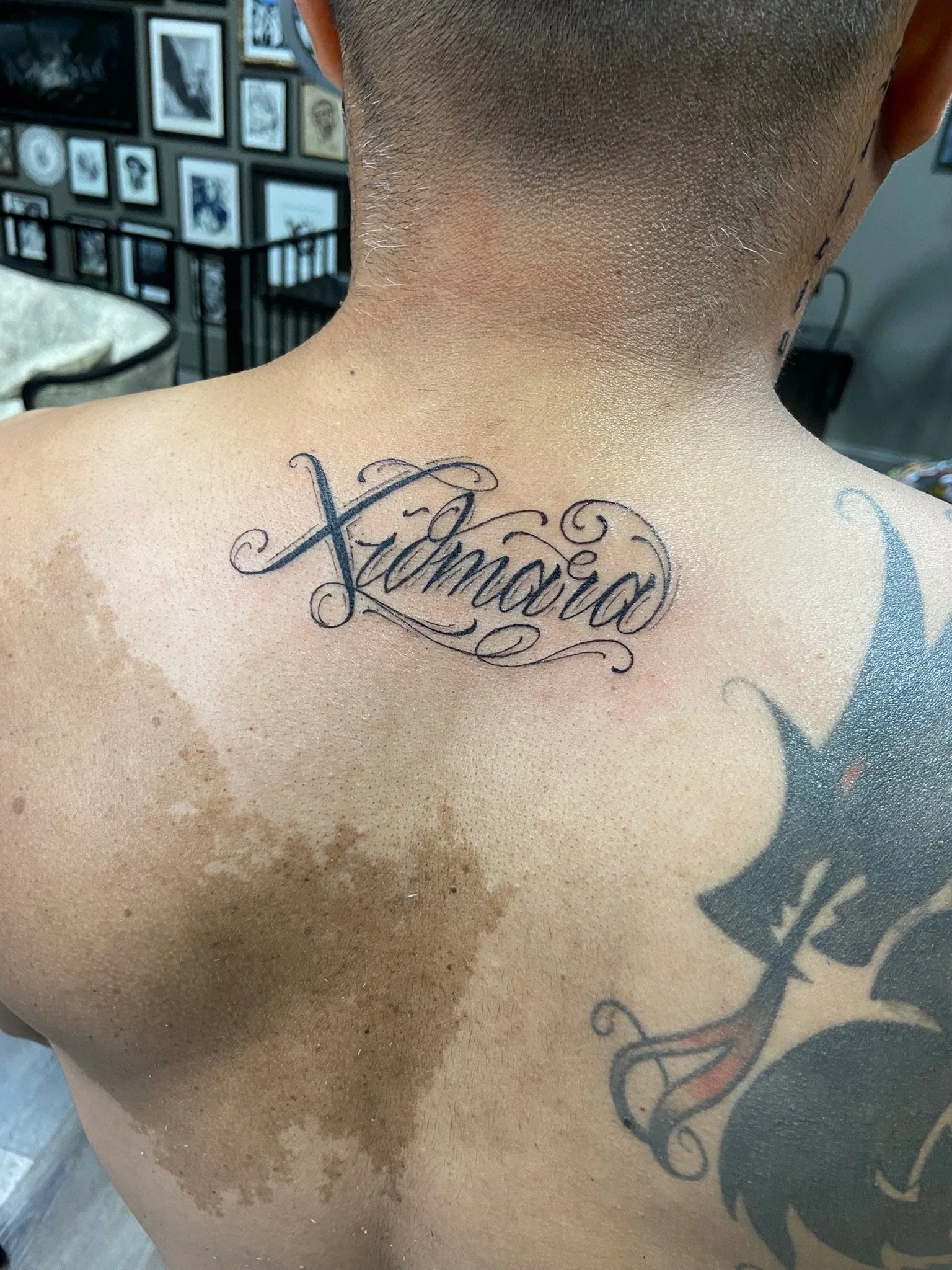 A close-up of a person's upper back and neck with a tattoo reading 'Xhavior' in cursive script. The person has short hair and smooth skin. In the background, there is a wall with multiple framed black-and-white and color images.