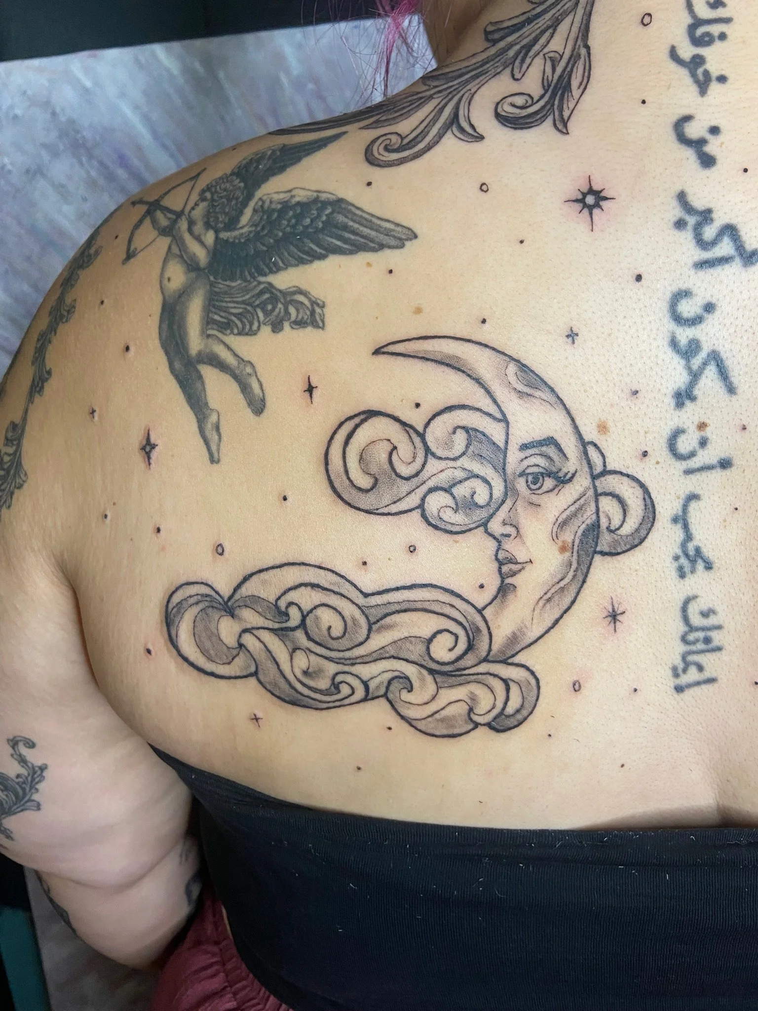 Tattoo of a detailed celestial scene featuring a moon with a woman's face, stars, and a mythical creature with wings and a bow, on a person's chest and shoulder.