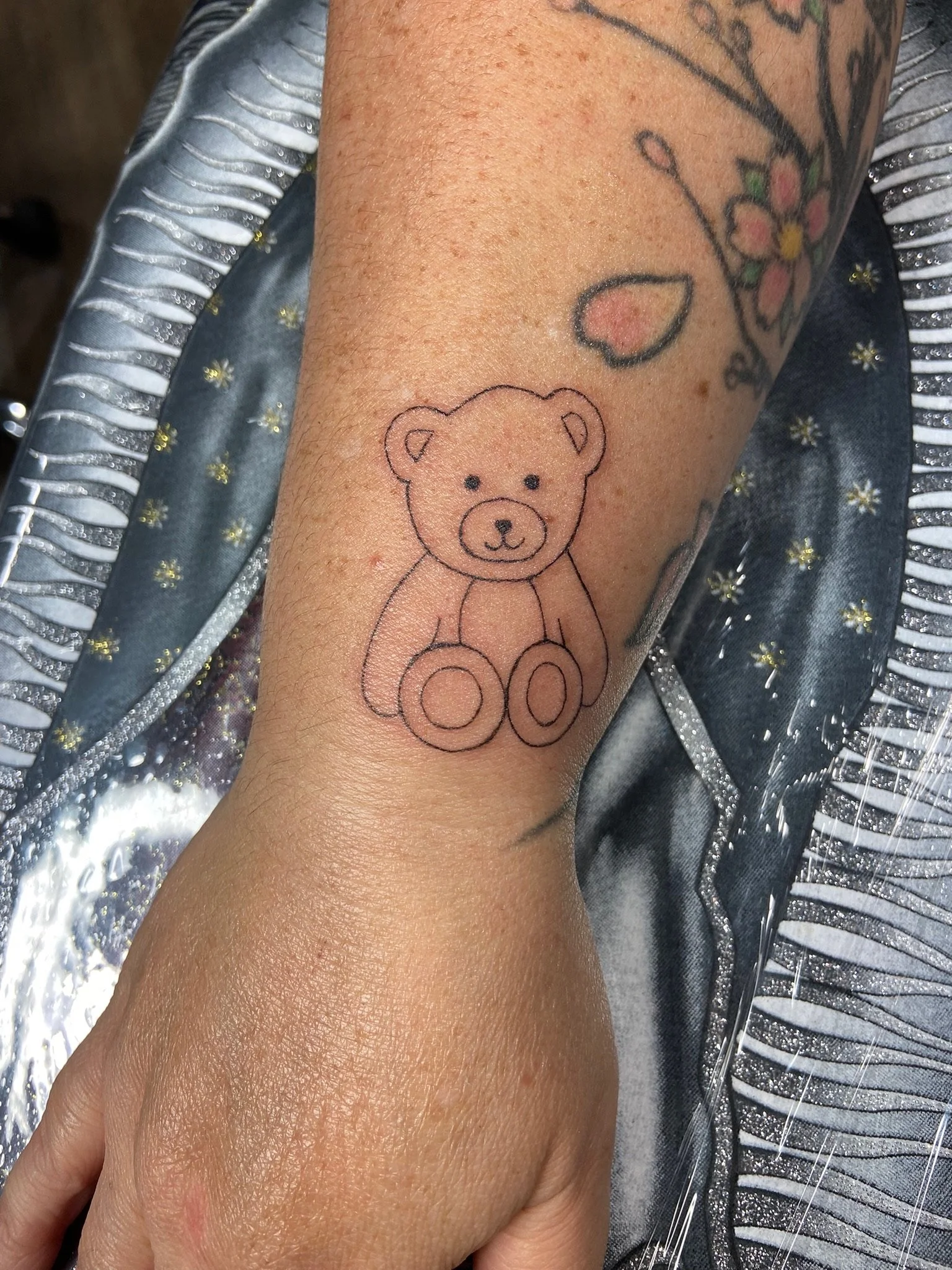 A tattoo of a teddy bear's face on a person's inner forearm, with partial tattoos of flowers and leaves above it. The person is dressed in shiny, striped, silver and blue clothing.