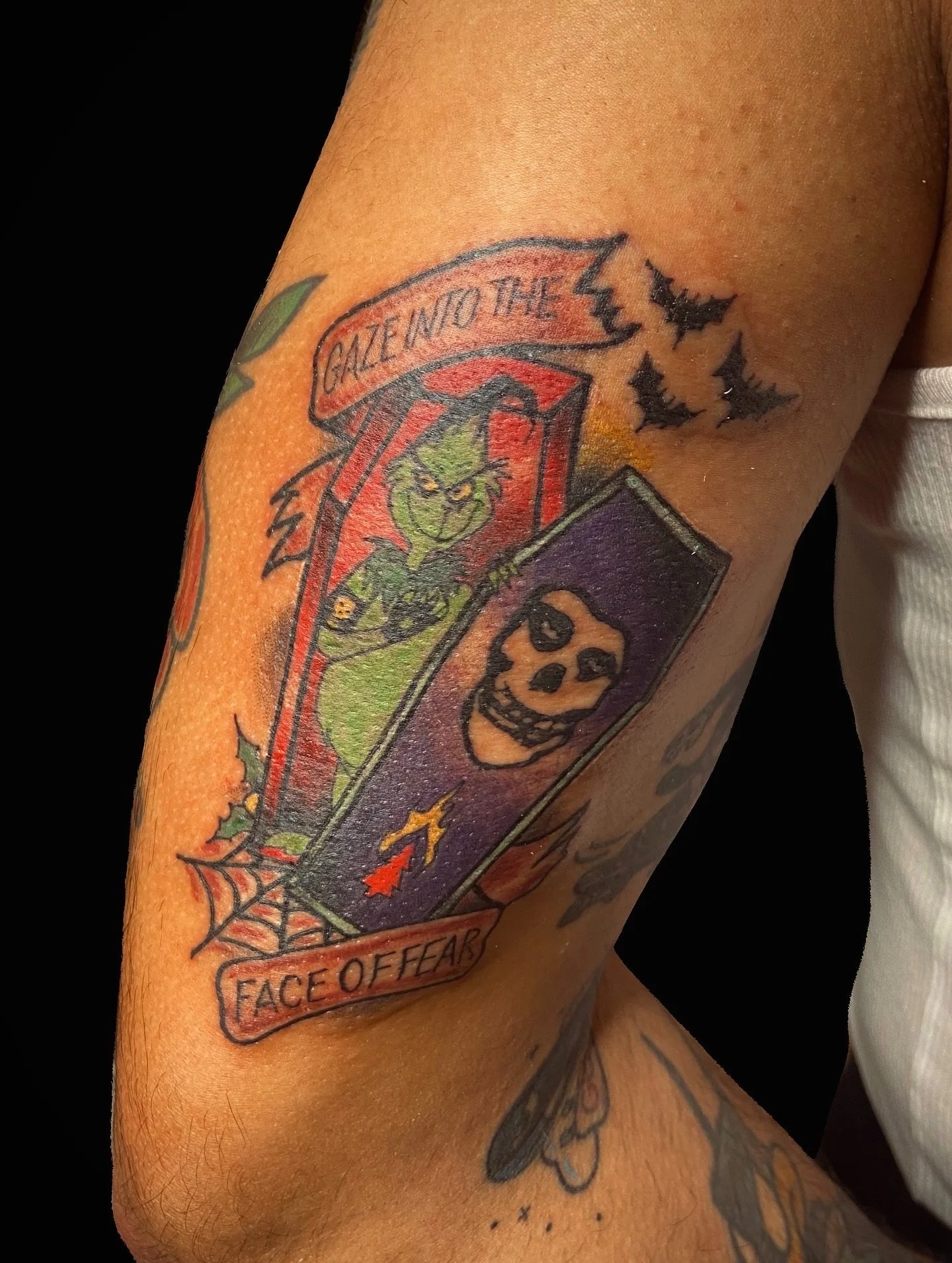 Tattoo on person's arm featuring a cartoonish green face with horns and a mischievous expression, with text 'GALLEITO THE' above it and a banner with 'FACE OF FEAR' underneath. The background includes bats and a spiderweb design.