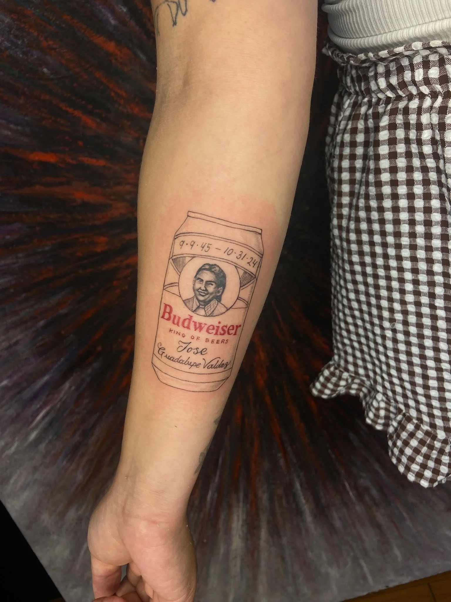 A person's arm with a tattoo of a Budweiser beer can featuring a portrait of a woman, with handwritten dates and names above and below the can on the tattoo.