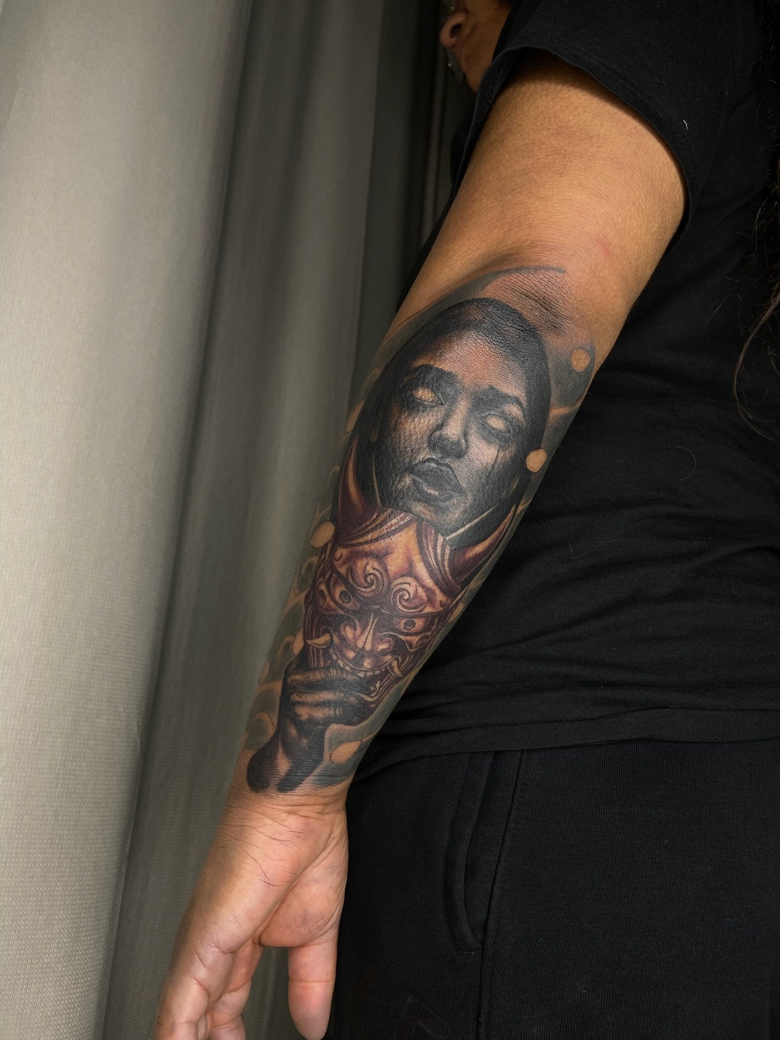 Close-up of a person's arm with a detailed black and gray tattoo of a woman's face and celestial elements, resting on another person's hand.