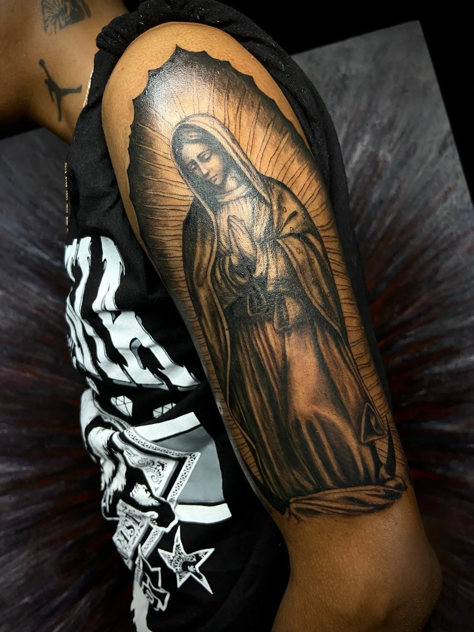 Tattoo of the Virgin Mary on a person's arm, with a halo of light behind her head, rendered in black and gray ink.
