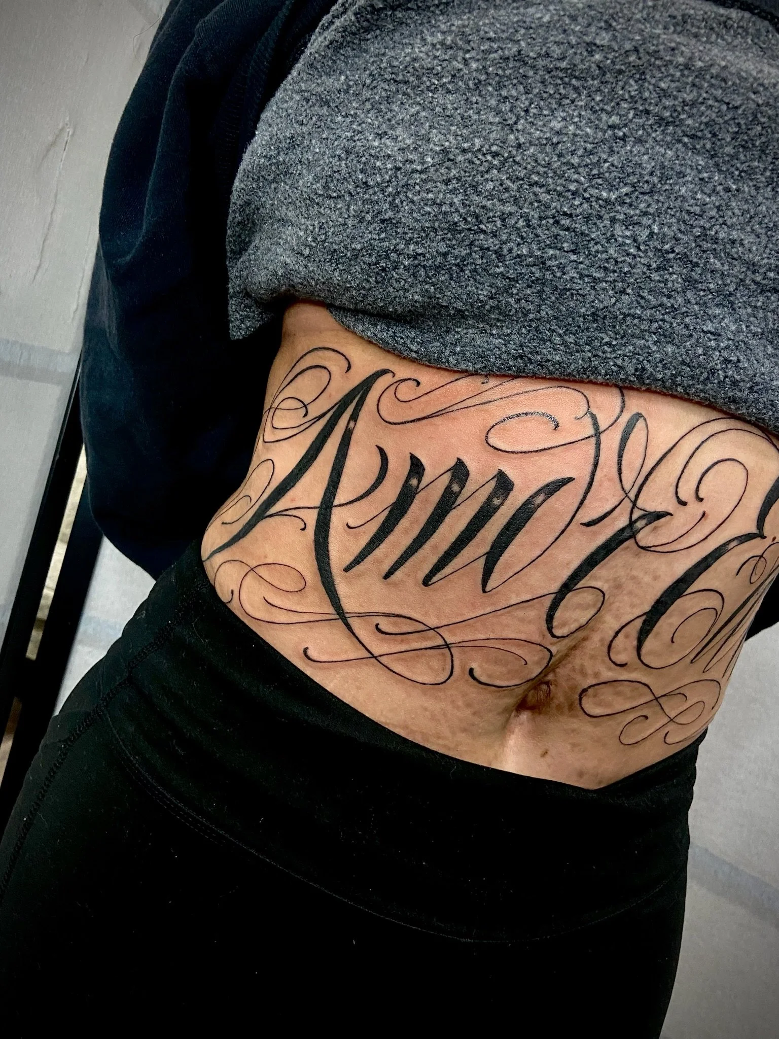 Close-up of a person's upper left arm with a large black tattoo featuring ornate cursive lettering.