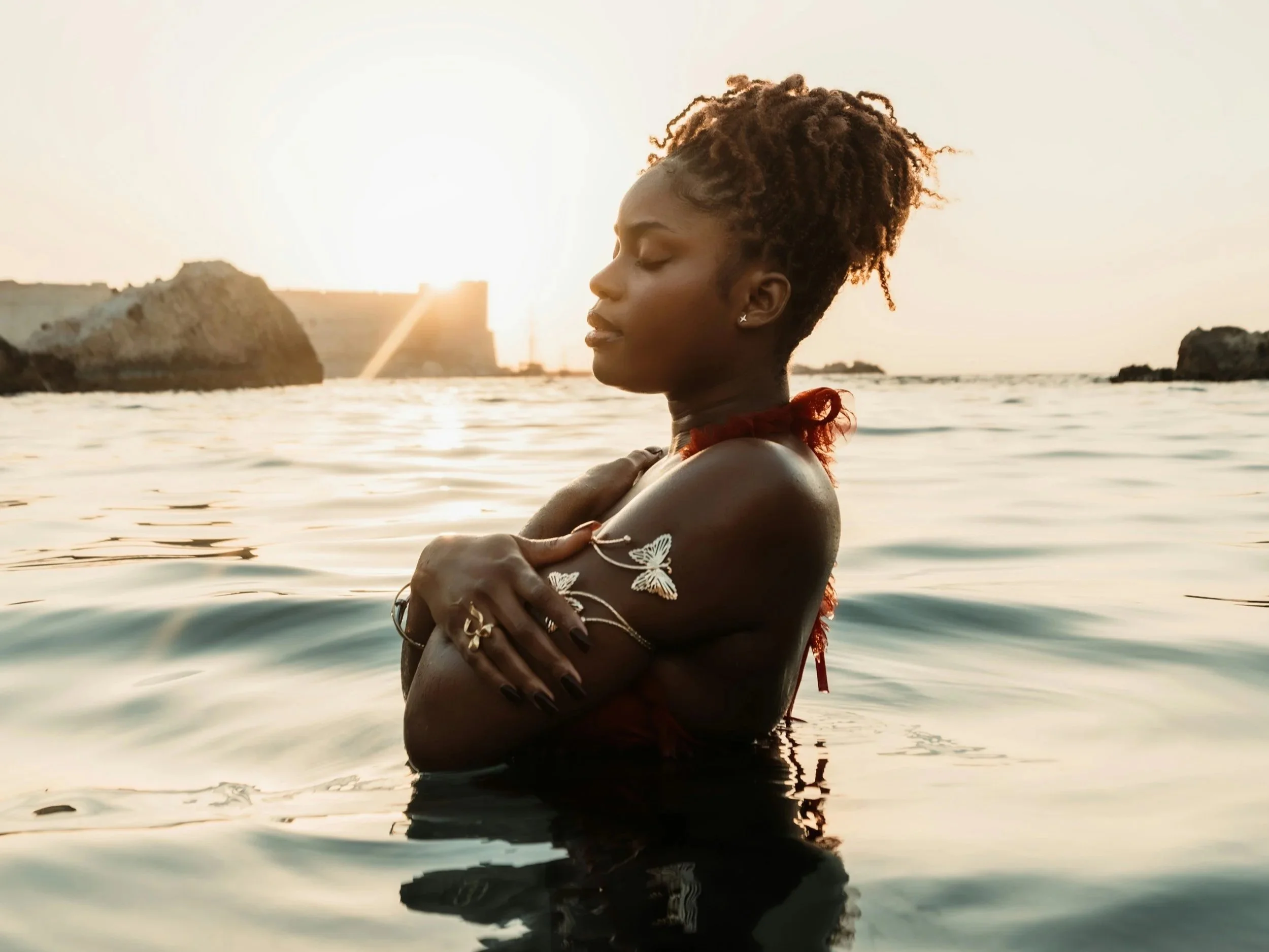 A woman with short curly hair and jewelry stands in the water with her eyes closed and arms crossed over her chest at sunset, with rocks and buildings in the background.