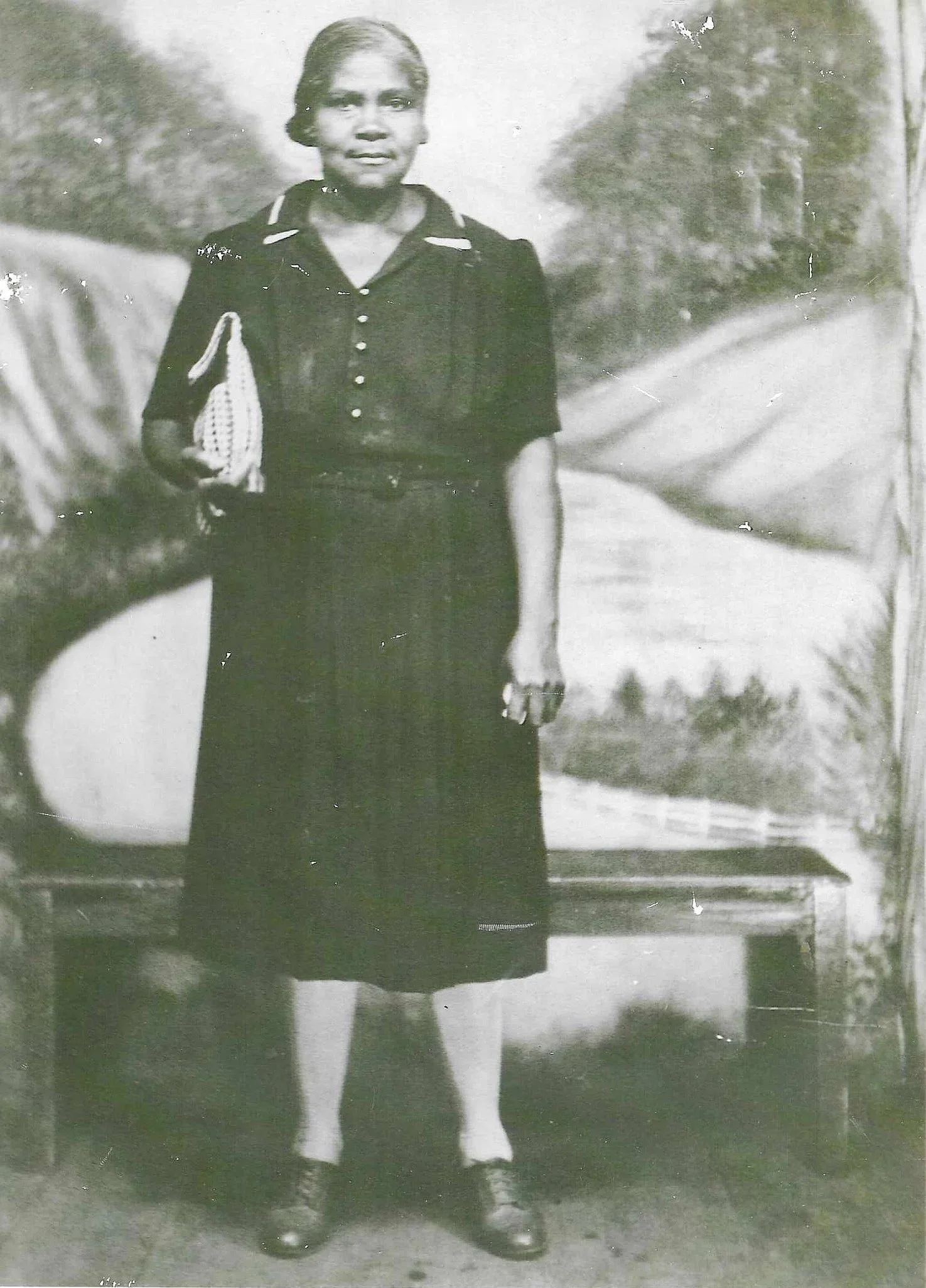 A young girl standing outdoors in a park or garden with trees in the background. She is wearing a dark dress with buttons down the front, a collared shirt underneath, and holding a small item or bag in her right hand.