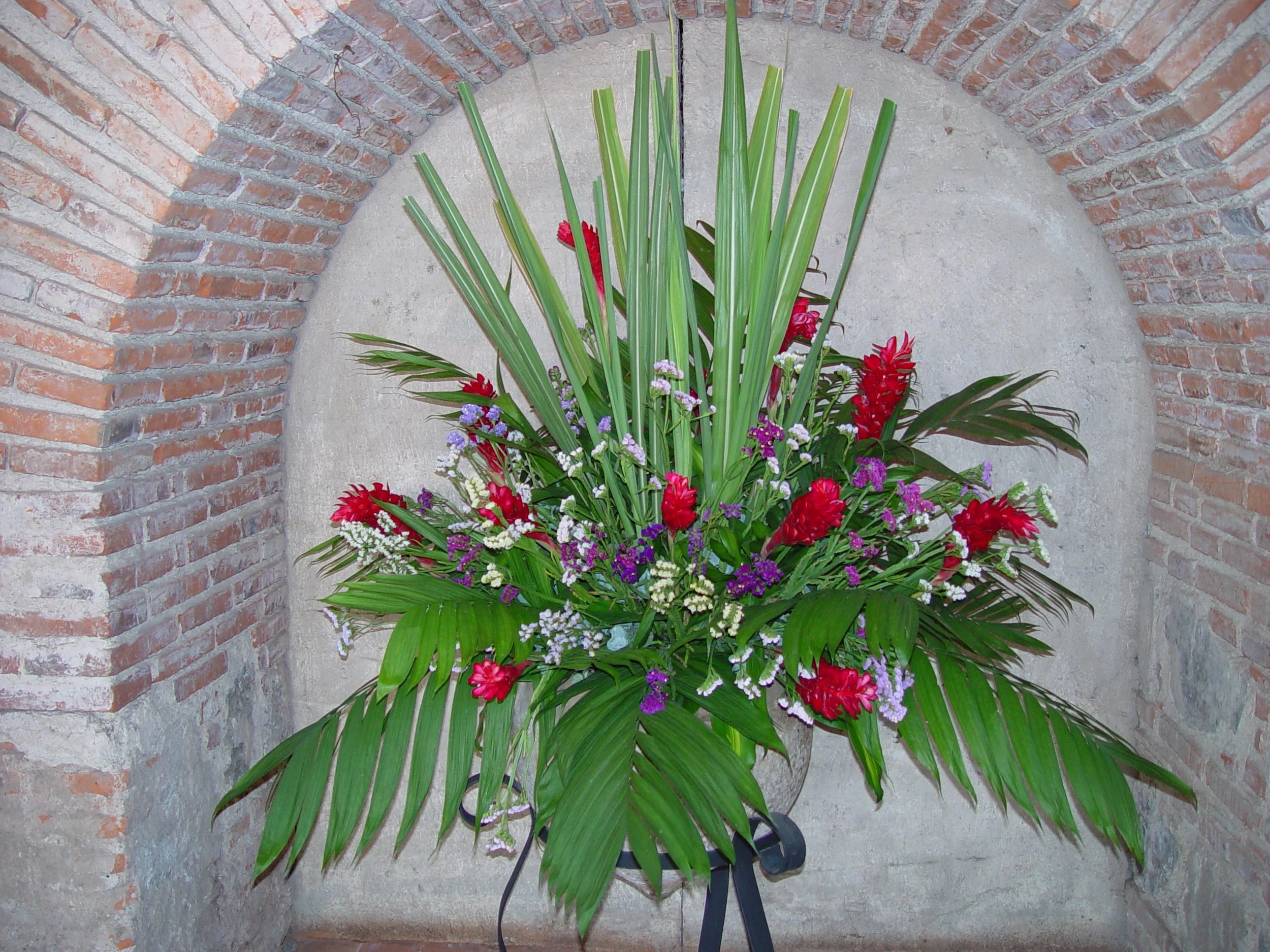 A bouquet of various flowers with red, purple, and white blossoms, and long green leaves, placed on a stand against a brick wall and concrete background.