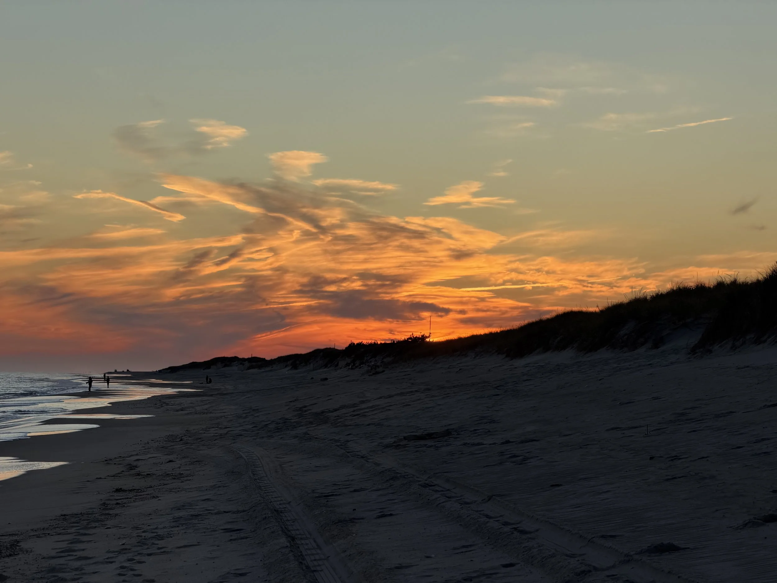A sunset over a beach with a colorful sky, sand dunes, and a few people walking along the shoreline.