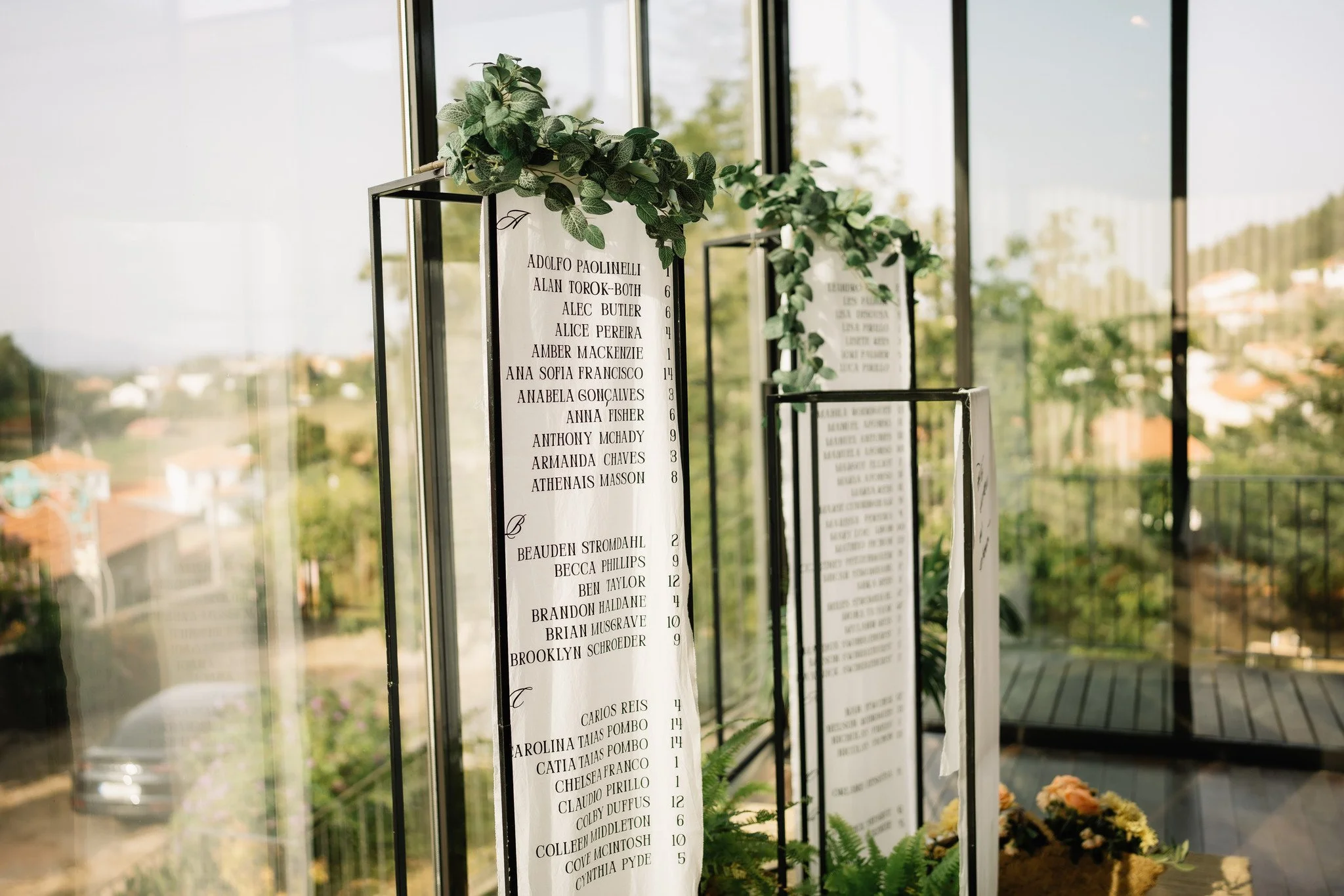 Elegant outdoor wedding setup with a round table draped in cream fabric, decorated with a large floral bouquet, framed guest sign, and vintage white rotary phone. A tall mirror with gold ornate top reflects the scene. In the background, guests gather under a white umbrella amidst greenery.