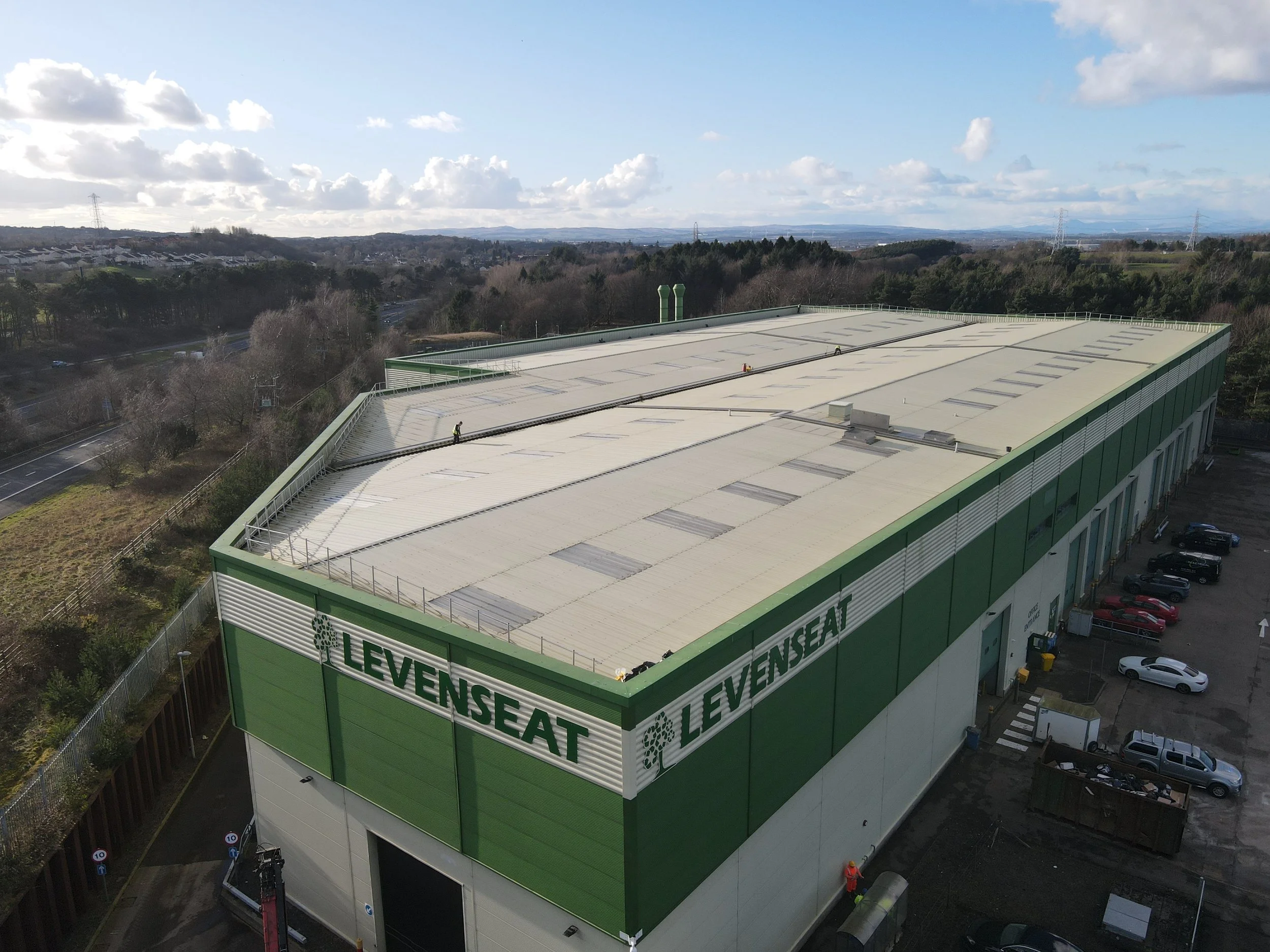 A large industrial building with green and white exterior, with the words 'Levenset' written on the side, situated in a semi-rural area with trees, roads, and parking lot nearby. The roof has multiple vents and skylights, and there are workers on the roof.