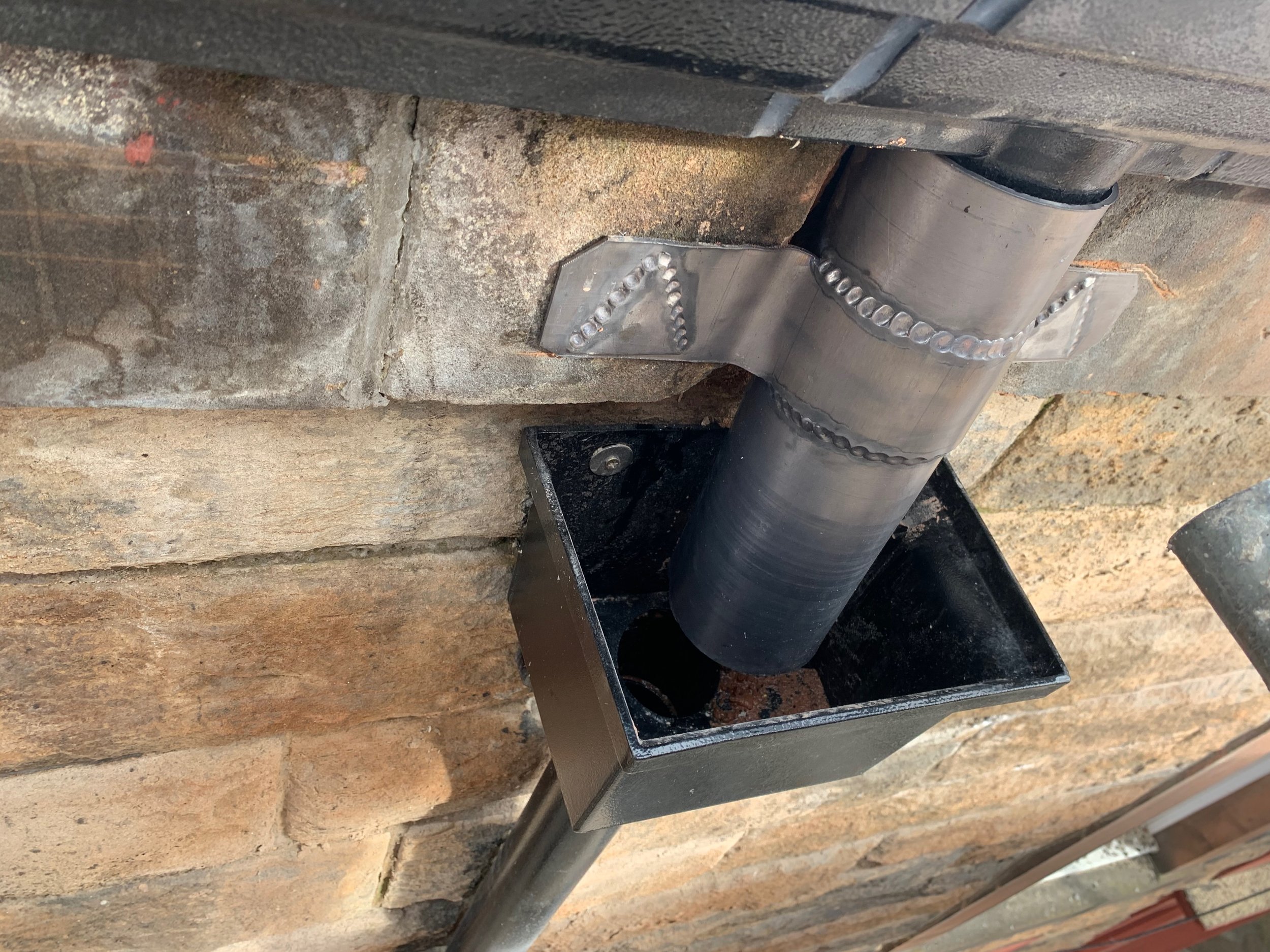 Close-up of a metal pipe with welds, mounted underneath a black metal frame, next to a concrete or stone wall.