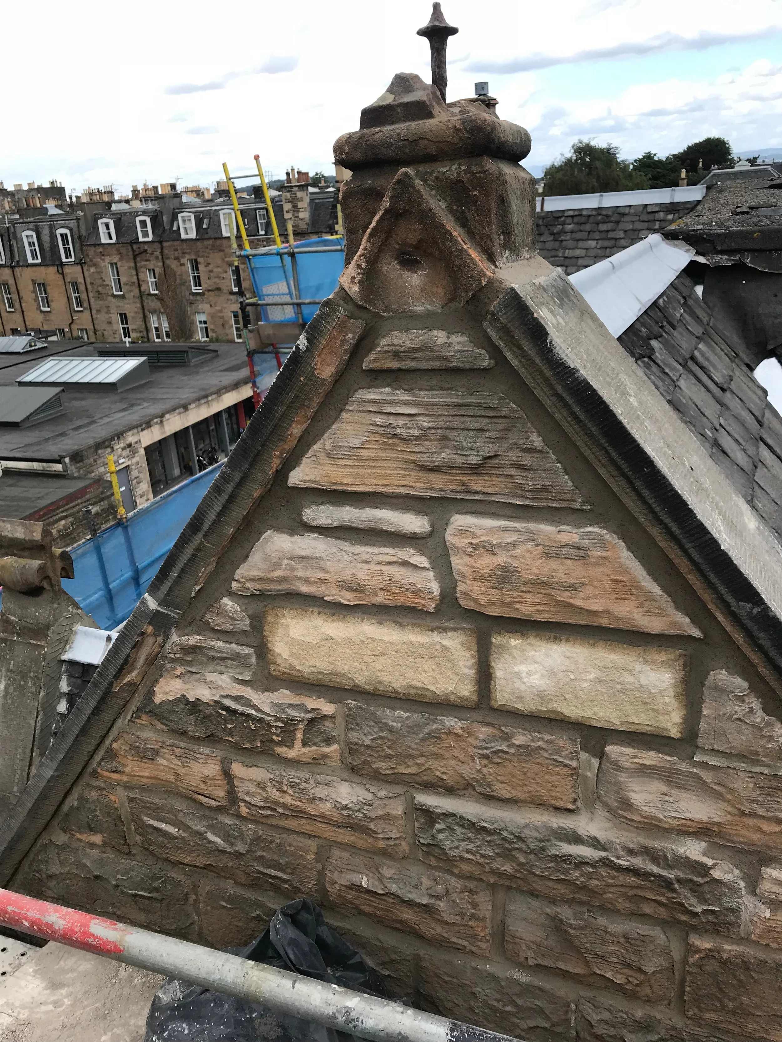 Close-up of a chimney on a roof under construction or repair, with bricks and mortar visible, and a background of neighboring rooftops and cloudy sky.