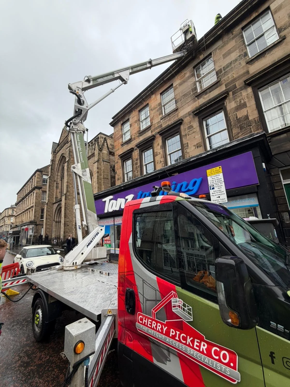 A cherry picker lift truck with an extended arm is working on the upper part of a multi-story building with stone facade and windows. The truck has a red and green body with logo and text, and is positioned on a city street. In the background, a sign for a tanning salon and other buildings are visible.
