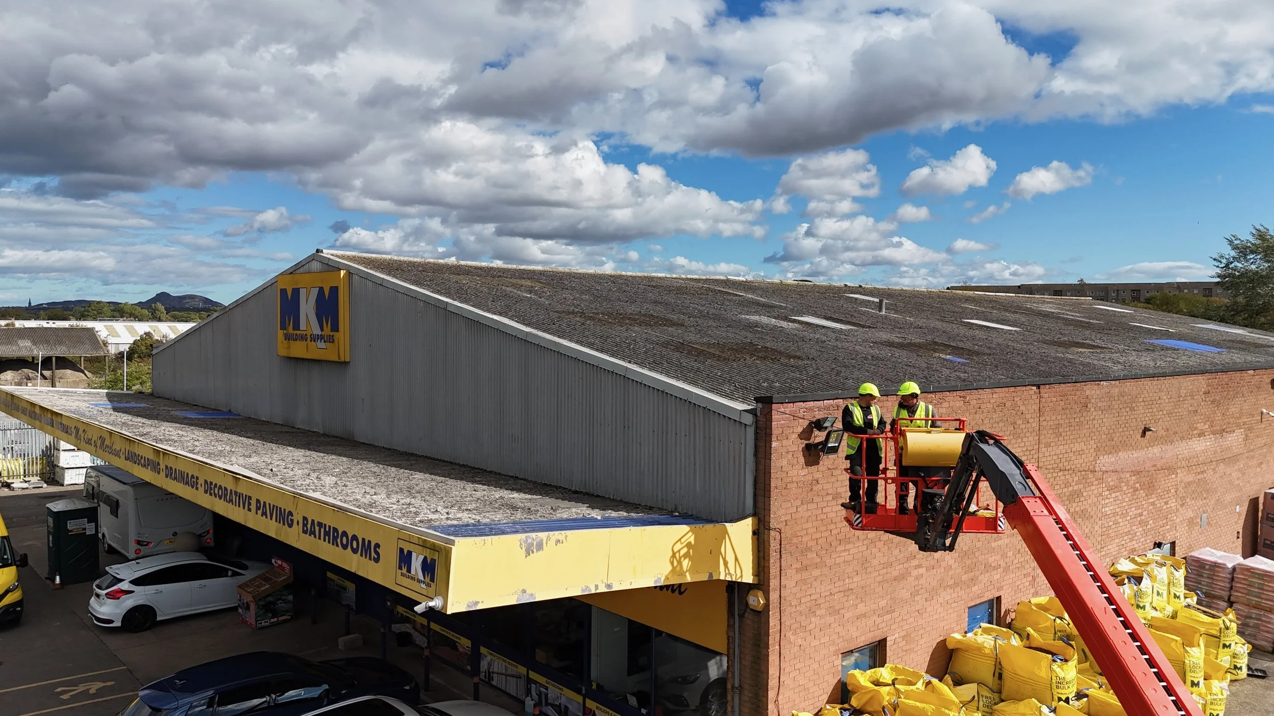 Two construction workers wearing safety helmets and vests operate a cherry picker lift next to a brick building. The background shows a warehouse with a sign reading 'MK Building Supplies' and a parking lot with several cars. The sky is partly cloudy.