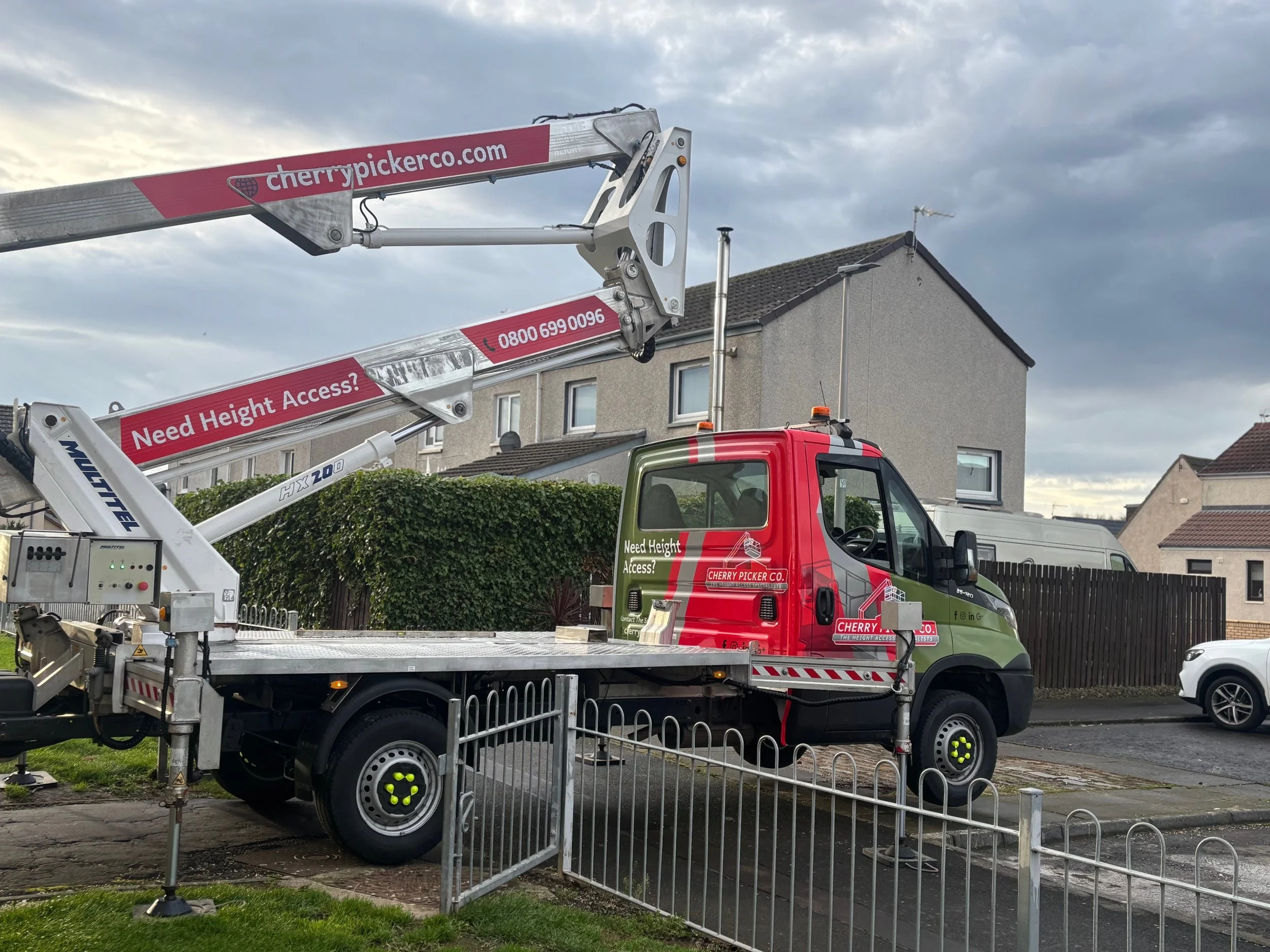 Mobile cherry picker lift truck with extended arm and platform, labeled with company contact information, positioned beside a house with a tiled roof, under a cloudy sky.