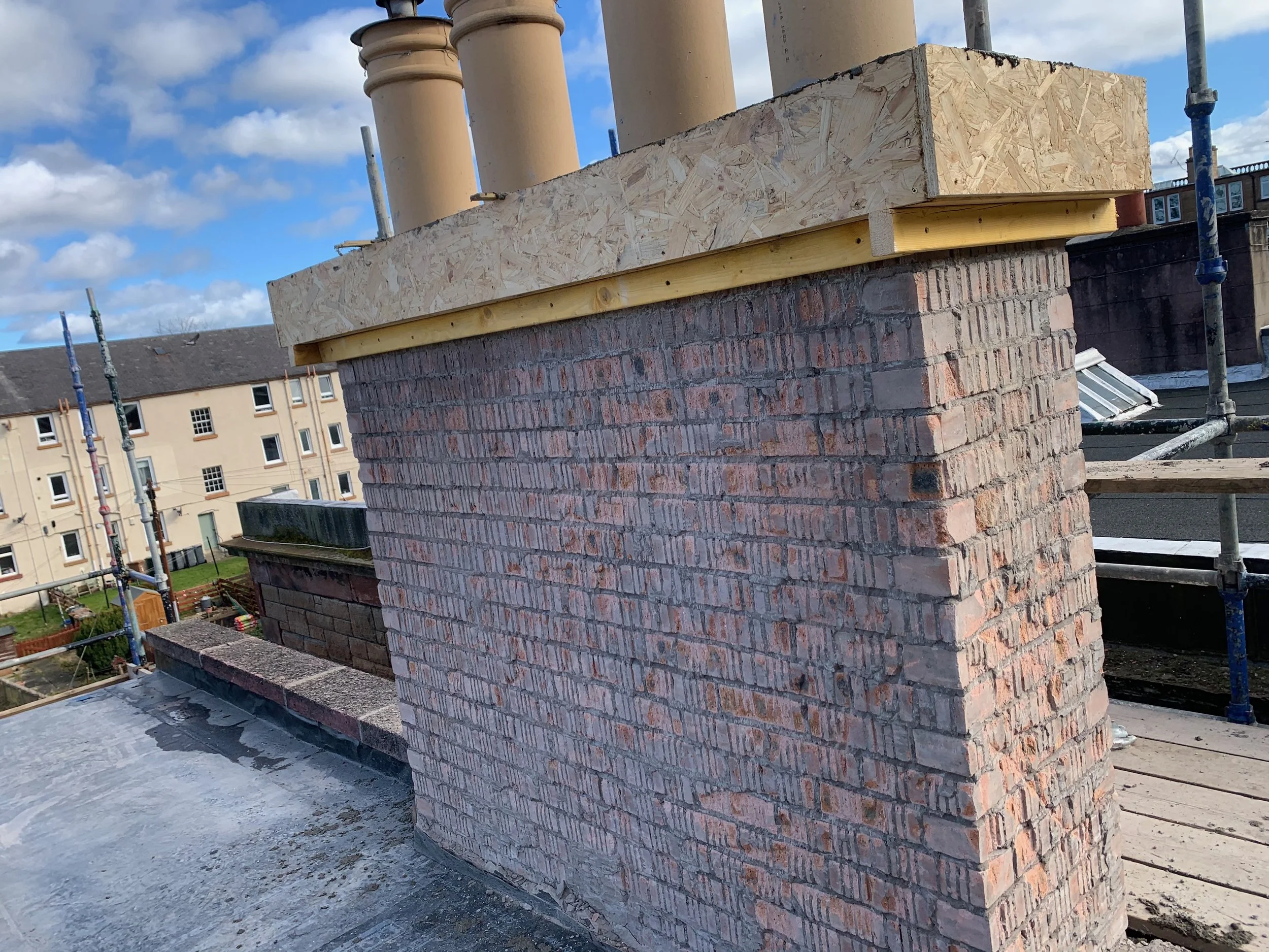 Construction site showing a brick chimney with scaffolding around it, and a new brick work layer above it under a partly cloudy sky.