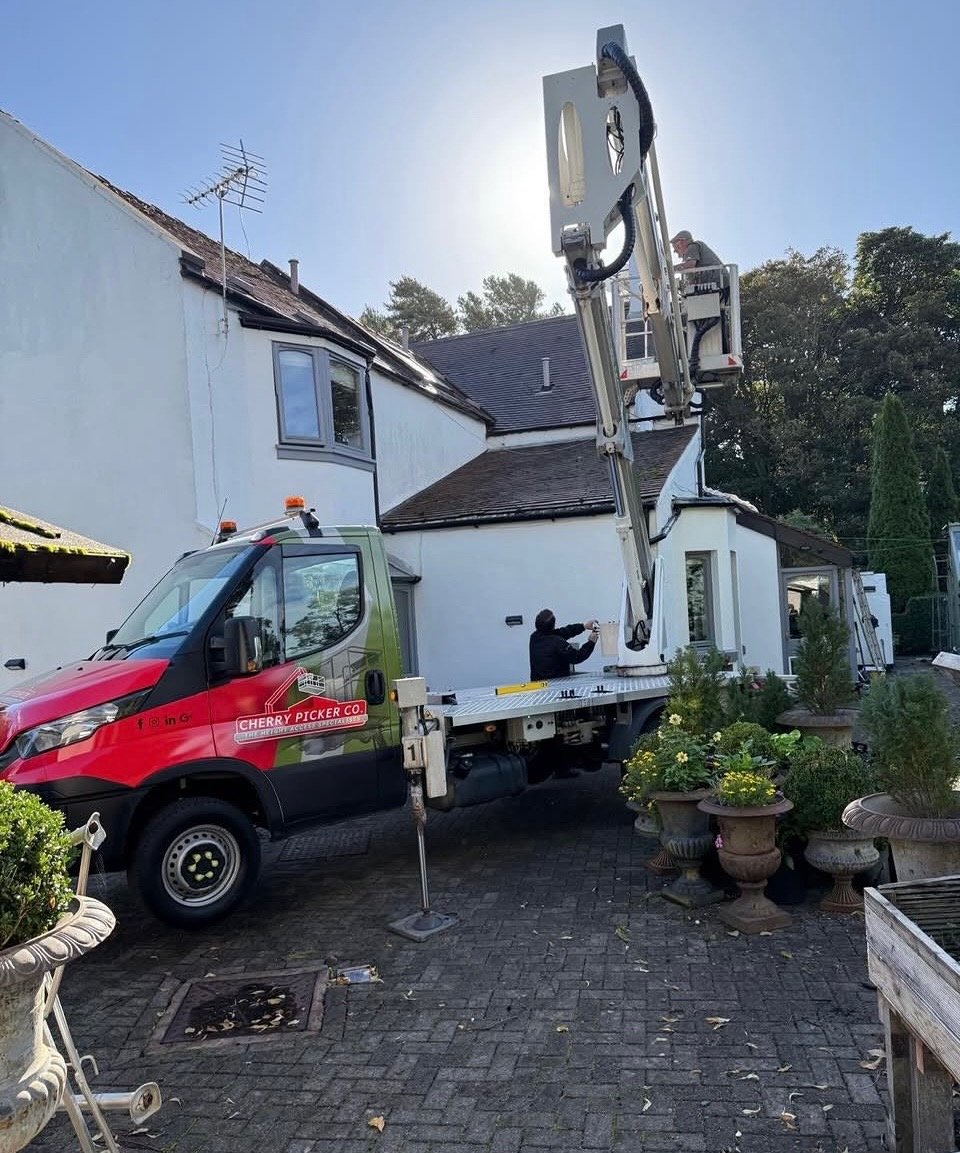 A cherry picker truck positioned in a residential driveway with a worker in the elevated bucket performing maintenance or repairs on a house roof.