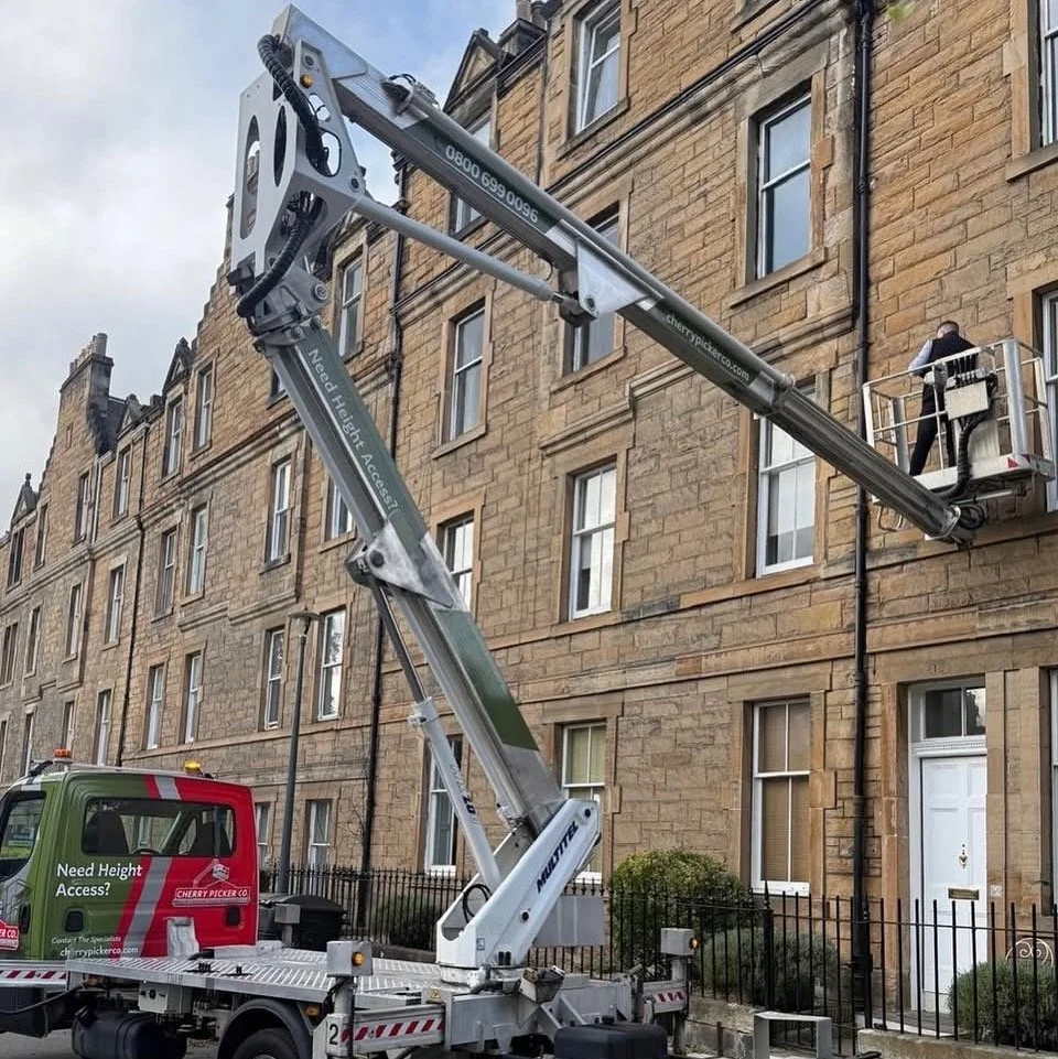 A worker using a cherry picker elevated at a brown brick apartment building to perform maintenance or repair work on the windows.