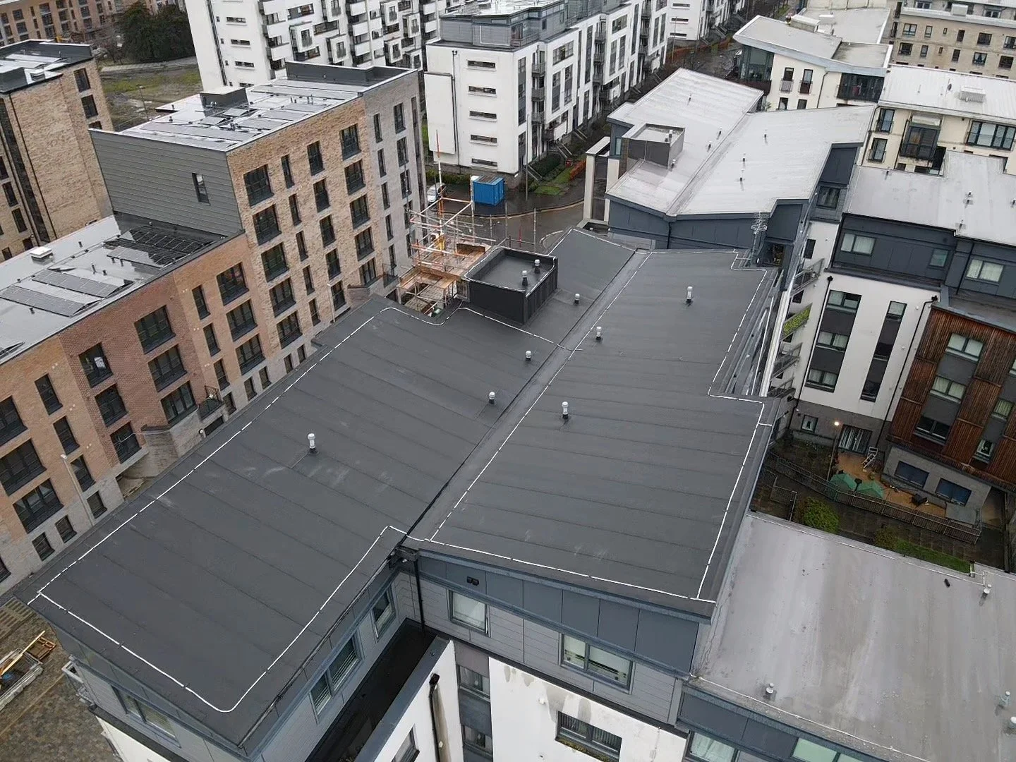 Aerial view of rooftops in an urban area, showing mostly flat roofs, some with solar panels, and surrounding high-rise buildings.