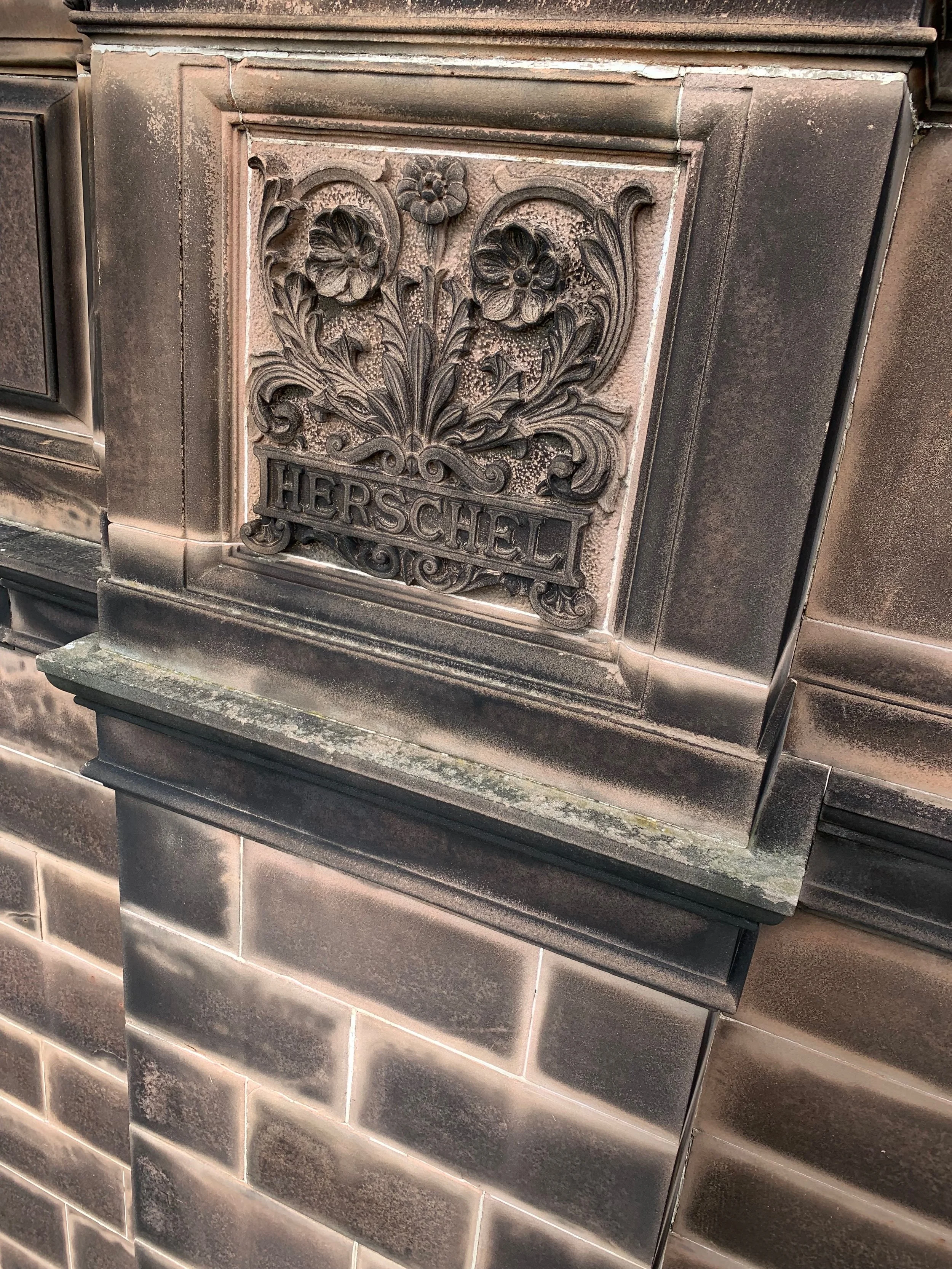 Close-up of an ornate, vintage metal plaque on a building, featuring floral and scroll designs with the word 'HERSCHEL' engraved at the bottom.