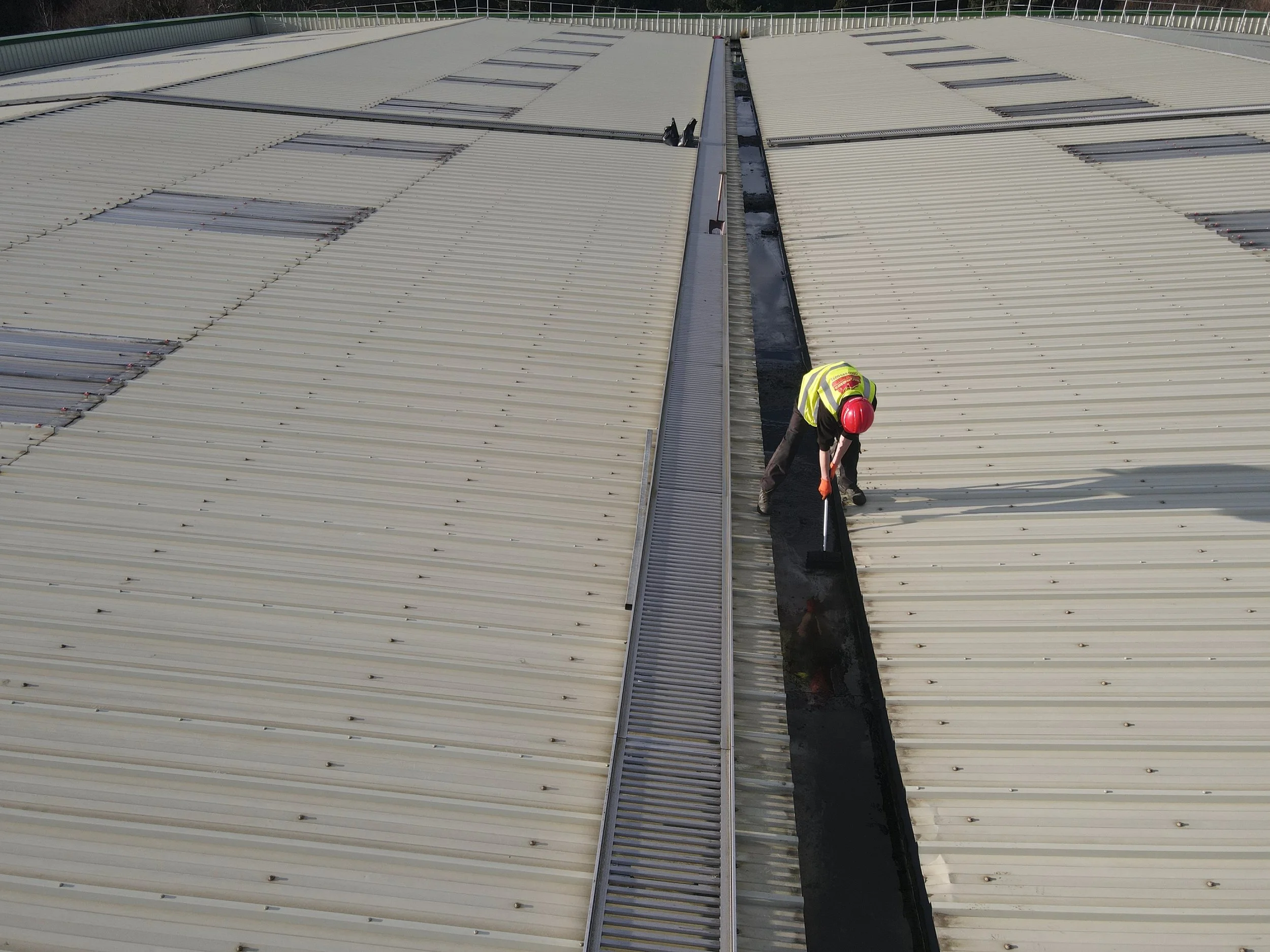 Worker in safety gear inspecting the roof of a large industrial building with a metal roof.