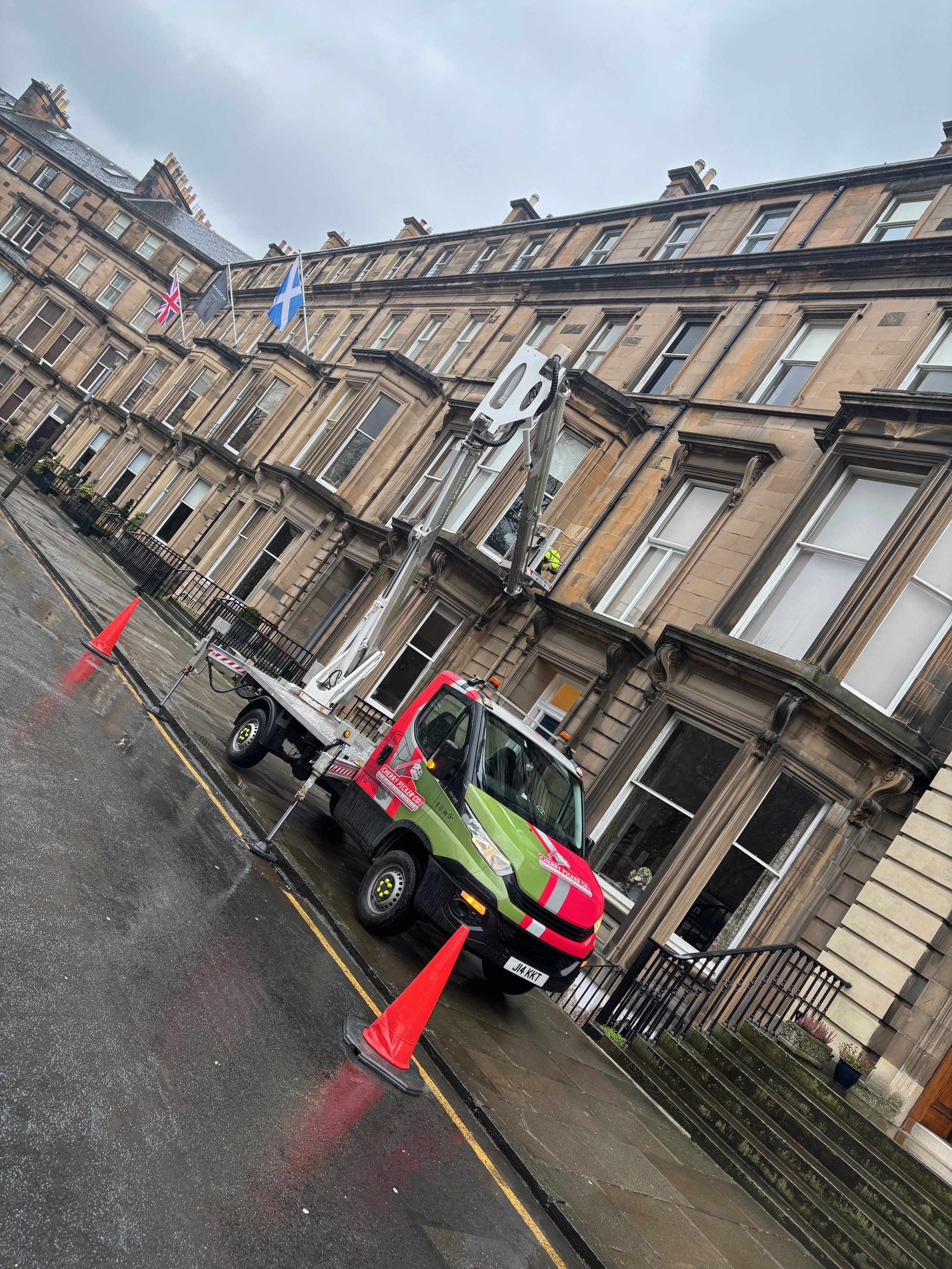 A residential street scene with an emergency vehicle parked in front of an apartment building. The vehicle is green with red markings and has a lift extended to work on the building's exterior. Orange cones are placed around the vehicle, and a worker is on the lift near the building's facade, which features several windows and flags, including the United Kingdom and Scottish flags, flying above the building.