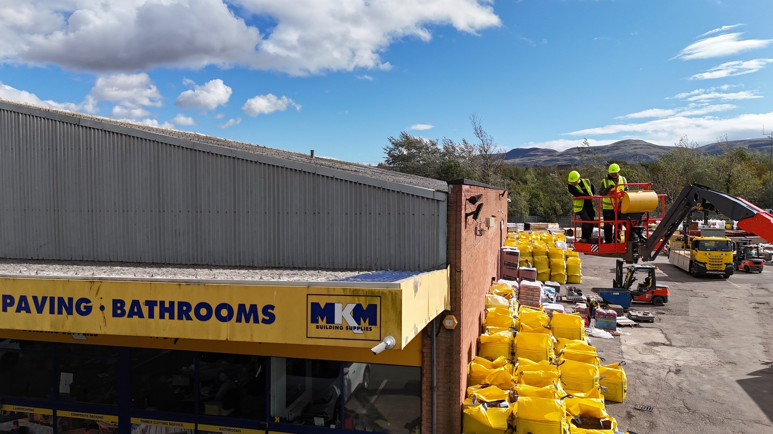 Two workers in safety vests and helmets operate an orange cherry picker lift, working near a brick building and a large sign for a store selling paving and bathrooms. The area is filled with yellow bags, construction materials, and equipment, with mountains and a mostly cloudy sky in the background.