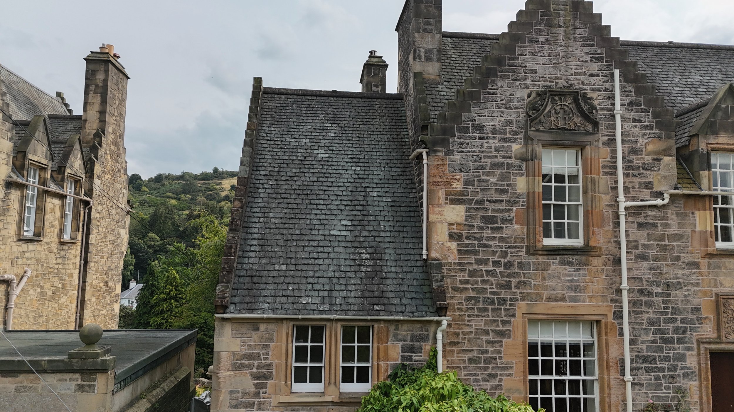 Close-up of historic stone buildings with slate rooftops and decorative stonework, including windows with white frames and greenery at the bottom, with a hill covered in trees in the background.