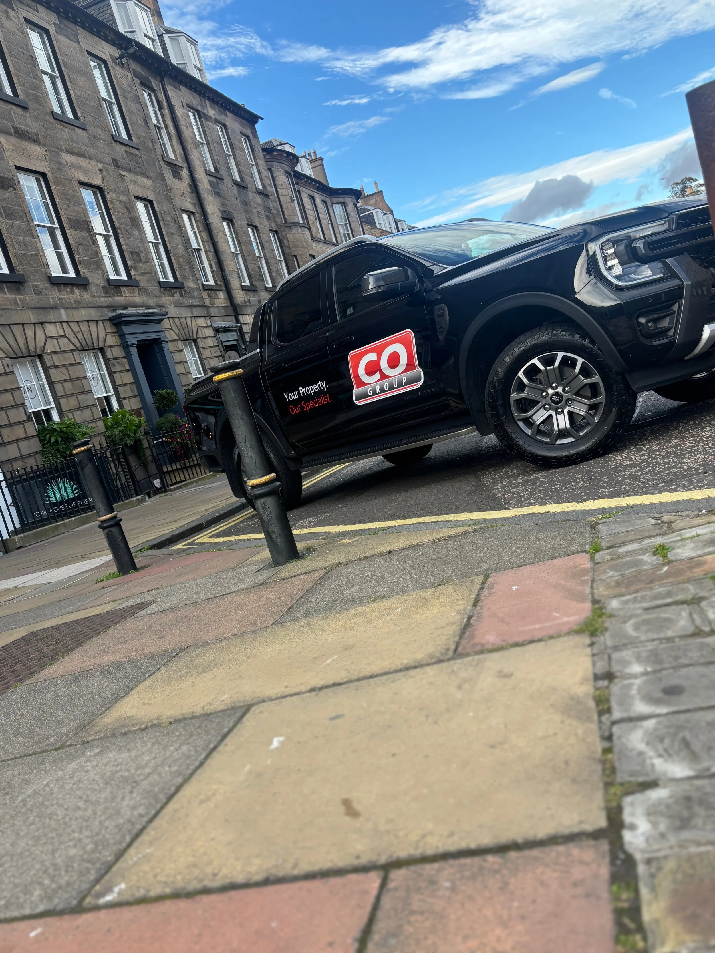 A black pickup truck with a red and white advertisement on the side, parked on a city street in front of old stone buildings with multiple windows. The street has a sidewalk with a multicolored tile pattern, and various street fixtures including barriers and poles.